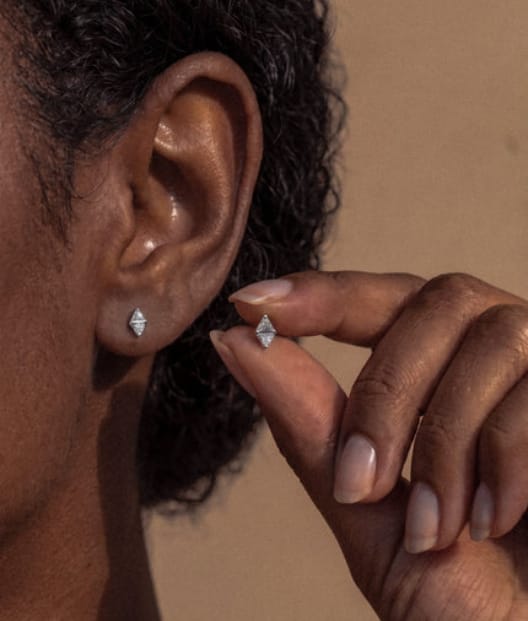 Una mujer de cabello castaño claro, con una camiseta blanca sin mangas, sonríe con dulzura; lleva un pendiente triangular azul pequeño «Trinity CZ» de Titanio-Noble Art que resalta su piercing sobre un fondo ligeramente difuminado.