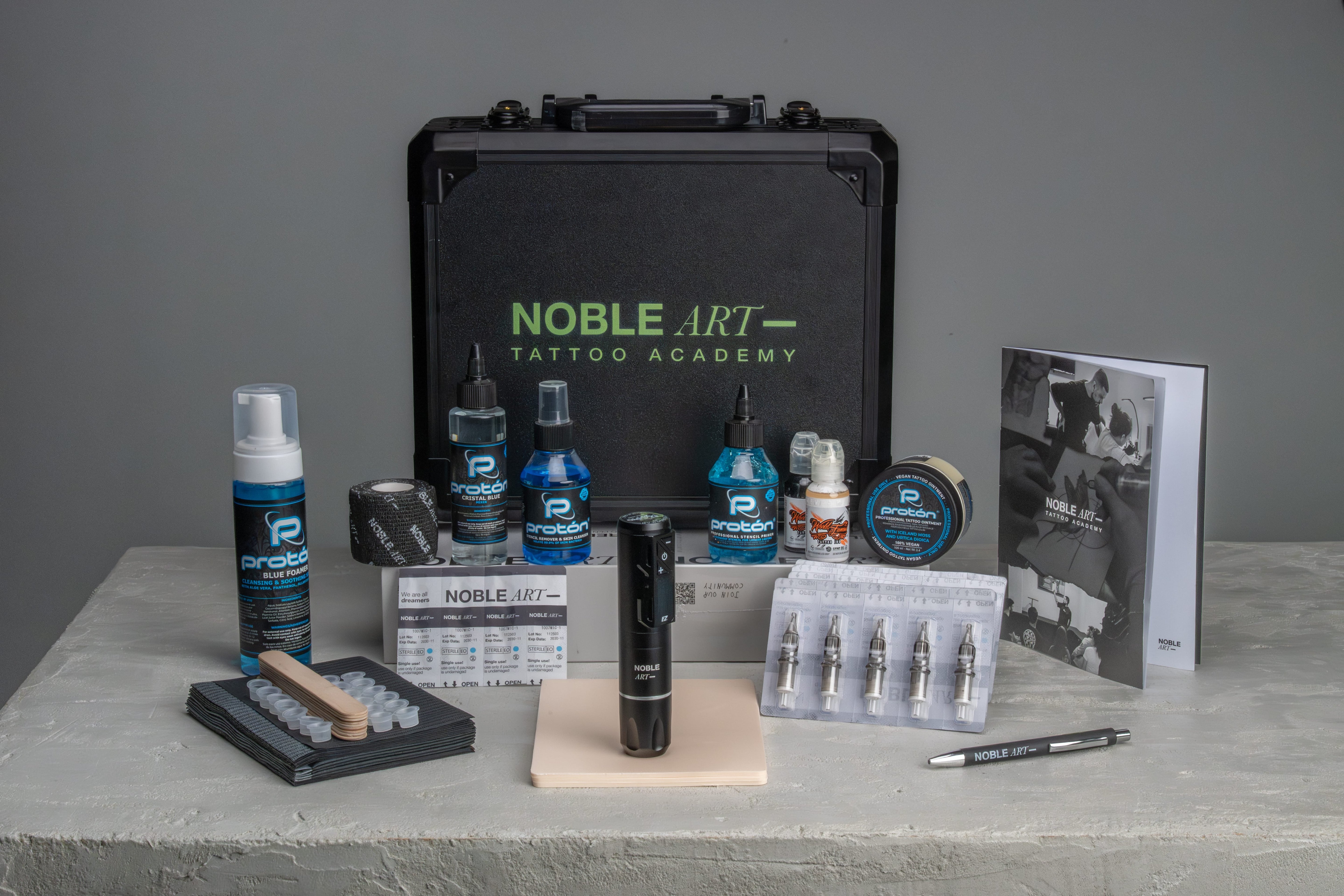 Kit de tatuaje con un estuche negro con la inscripción «Noble Art Tattoo Academy», una máquina de tatuar, frascos de tinta, agujas, cinta antideslizante, una guía de referencia y otros materiales para tatuar, todo ello sobre una superficie gris.