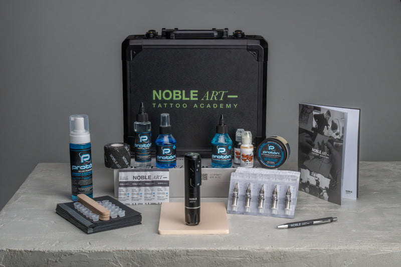 Kit de tatuaje con un estuche negro con la inscripción «Noble Art Tattoo Academy», una máquina de tatuar, frascos de tinta, agujas, cinta antideslizante, una guía de referencia y otros materiales para tatuar, todo ello sobre una superficie gris.