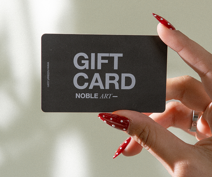 Una mano con las uñas pintadas de rojo a lunares sostiene una tarjeta regalo negra en la que se lee «GIFT CARD NOBLE ART» en letras blancas. La luz del sol proyecta suaves sombras sobre el fondo.