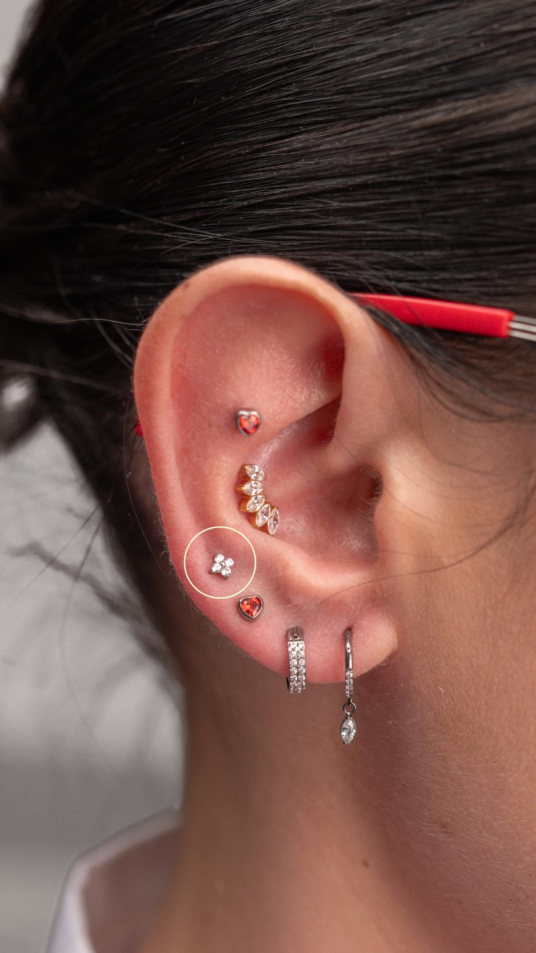 Un primer plano de una oreja en el que se aprecian varios piercings en el «lóbulo superior», realizados por NOBLE ART OFFICIAL COORP, con pendientes en forma de corazón, un pendiente floral, un aro con joyas y una patilla de gafas roja: una elegante muestra del estilo de Noble Art.