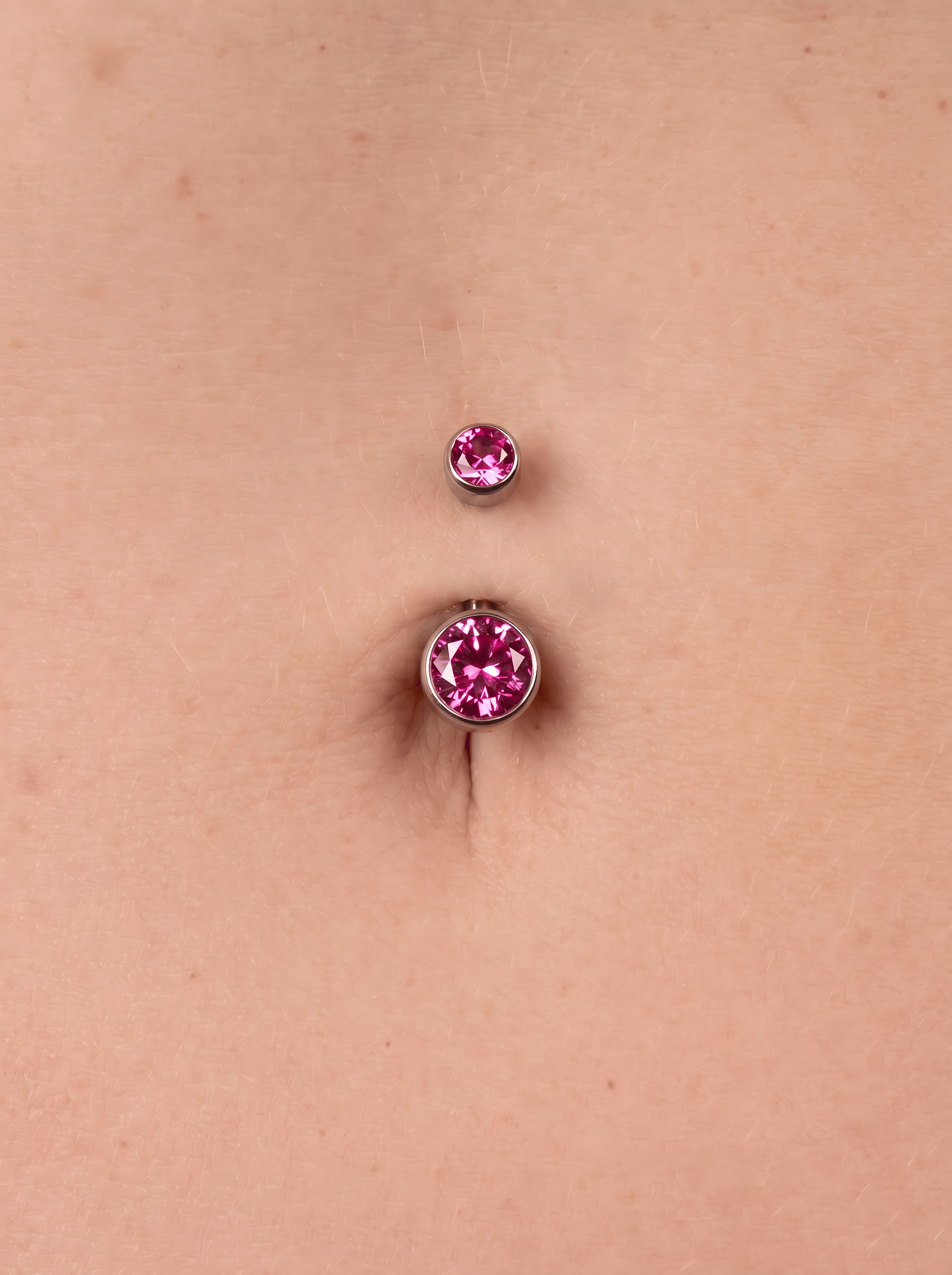 Un primer plano de un ombligo de piel clara adornado con el «Titanio- Noble Art OMBLIGO CZ ROSA», un piercing plateado con dos piedras preciosas de color rosa: una encima y otra dentro del ombligo.