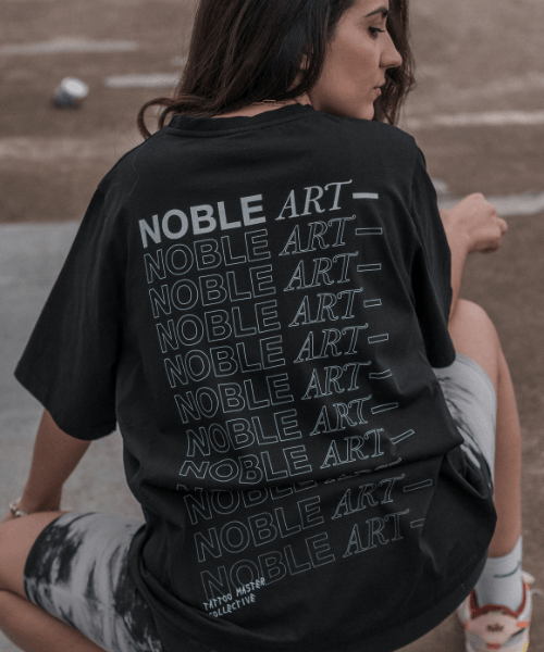 Una persona está sentada al aire libre, de espaldas, con una camiseta negra de Noble Art con el logotipo blanco repetido en la espalda, quizá esperando una eliminación láser o presumiendo de un nuevo piercing.