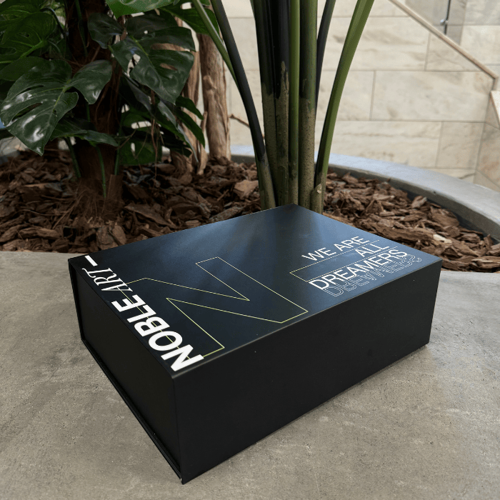 A black rectangular box with gold and white lettering reading "Noble Art" and "Beginner Tattoo Kit" rests on a concrete surface next to plants and mulch, capturing the creative spirit of Noble Art tattoo design.