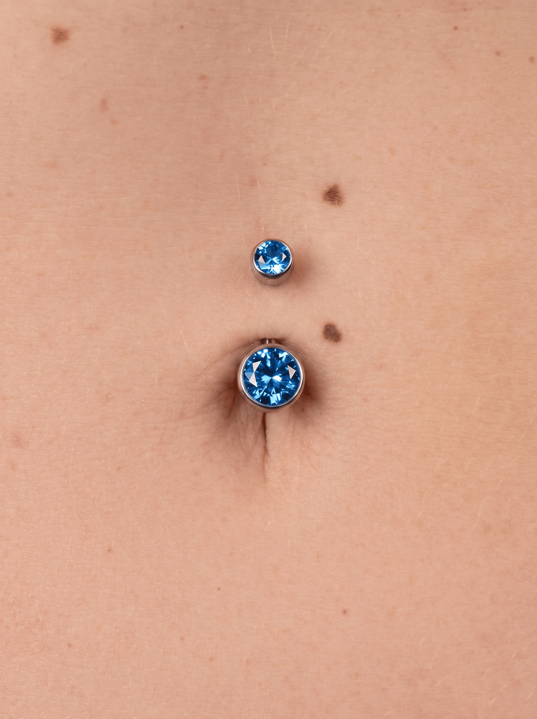 Close-up of a pierced navel, showing the “Blue CZ Navel” model by Titanio-Noble Art: two blue CZ stones with a silver finish placed above and inside the navel on fair skin.
