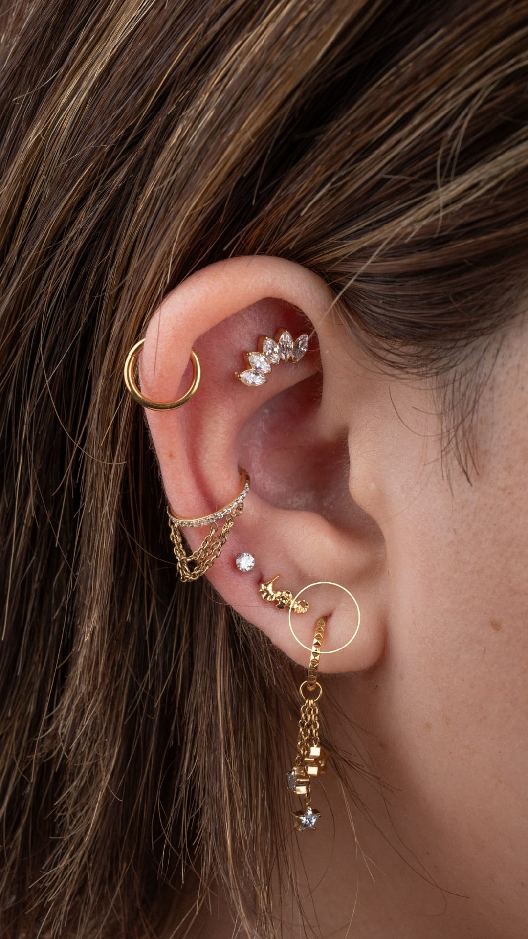Un primer plano destaca los originales piercings en las orejas de una persona, adornados con pendientes de oro de la marca Lóbulo —aros, pendientes de botón y cadenas con piedras preciosas de NOBLE ART OFFICIAL COORP—. Su cabello castaño claro con reflejos rubios encarna el espíritu de la autoexpresión.