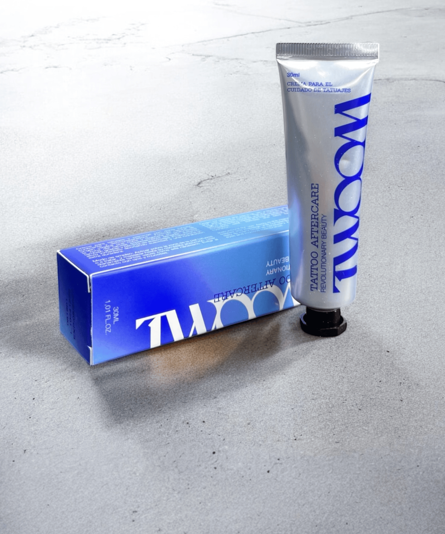 Un tubo de TWOOM Tattoo Aftercare, de NOBLE ART BEAUTY SOCIEDAD LIMITADA, se encuentra en posición vertical junto a su caja azul y blanca sobre una superficie gris claro; ambos presentan unas letras azules en negrita inspiradas en el Noble Art. Ideal para los amantes de los tatuajes y para quienes se plantean la eliminación con láser.
