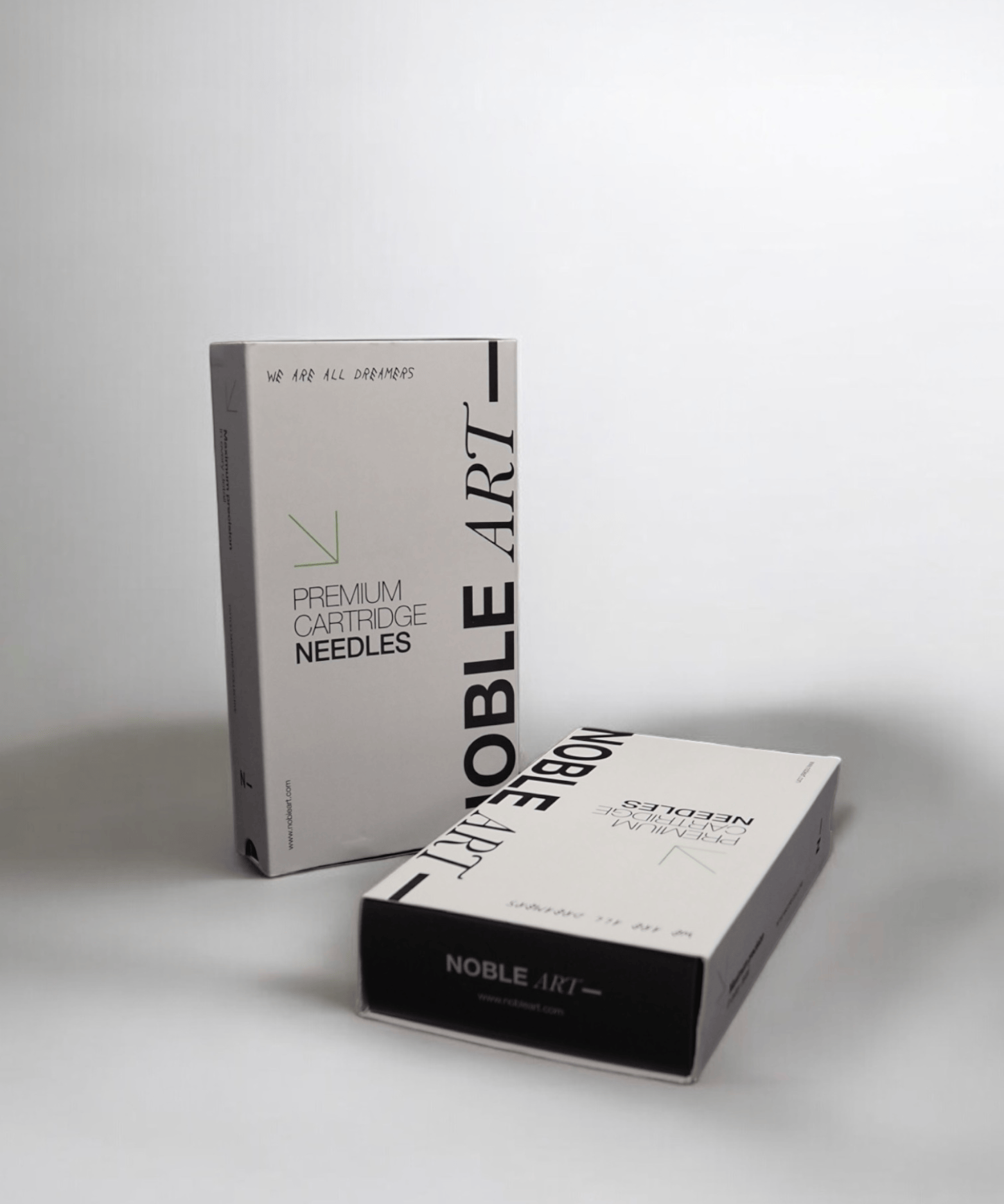 Se muestran dos cajas rectangulares con la etiqueta «Agujas round liner» de NOBLE ART BEAUTY SOCIEDAD LIMITADA, una en posición vertical y otra tumbada, ambas con un diseño minimalista en blanco y negro sobre un fondo blanco liso.