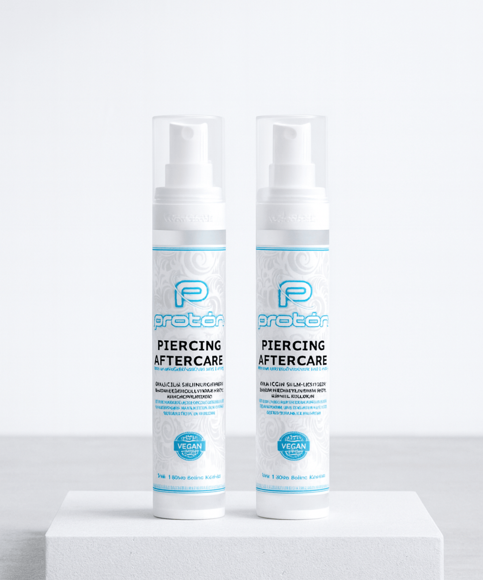 Dos frascos blancos de Aftercare Piercing Proton, de NOBLE ART BEAUTY SOCIEDAD LIMITADA, con etiquetas azules y plateadas, se muestran uno al lado del otro sobre una plataforma blanca con un fondo gris claro, ideales para el cuidado de tatuajes o piercings.