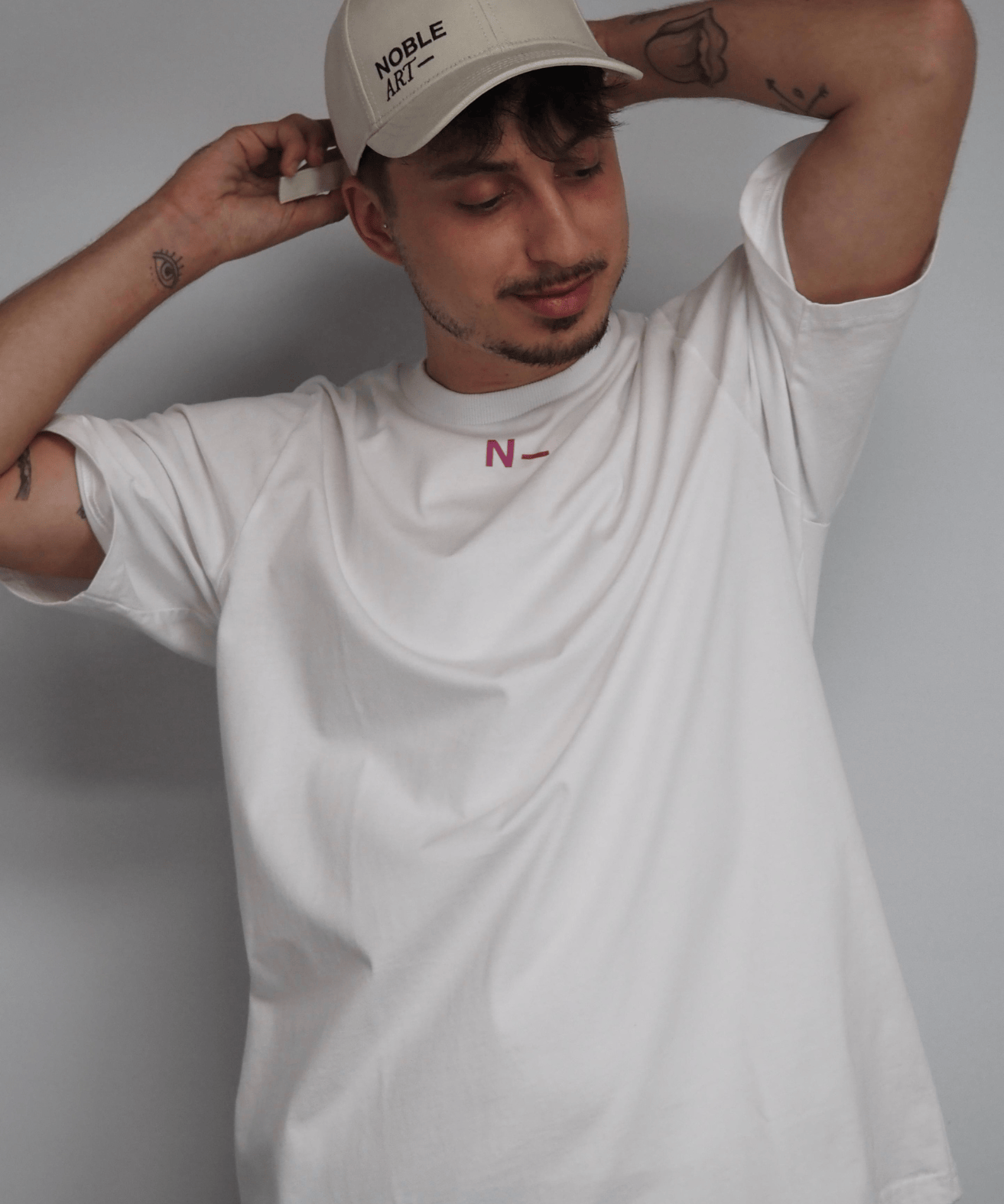 Una persona que lleva una gorra beige de Noble Art y una camiseta «pantera blanca» de Noble Art, con un pequeño emblema rojo en forma de «N», está de pie con los ojos cerrados y los brazos levantados, mostrando un discreto piercing mientras se toca la nuca.