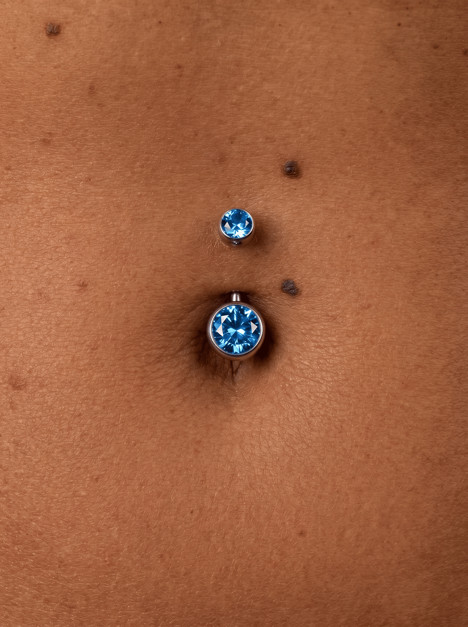 Eine Nahaufnahme des „Ombligo cz azul“-Bauchnabelpiercings von Titanio-Noble Art: Ein silberner Barbell mit blauen CZ-Steinen vor brauner Haut, daneben sind zwei kleine Muttermale zu sehen.
