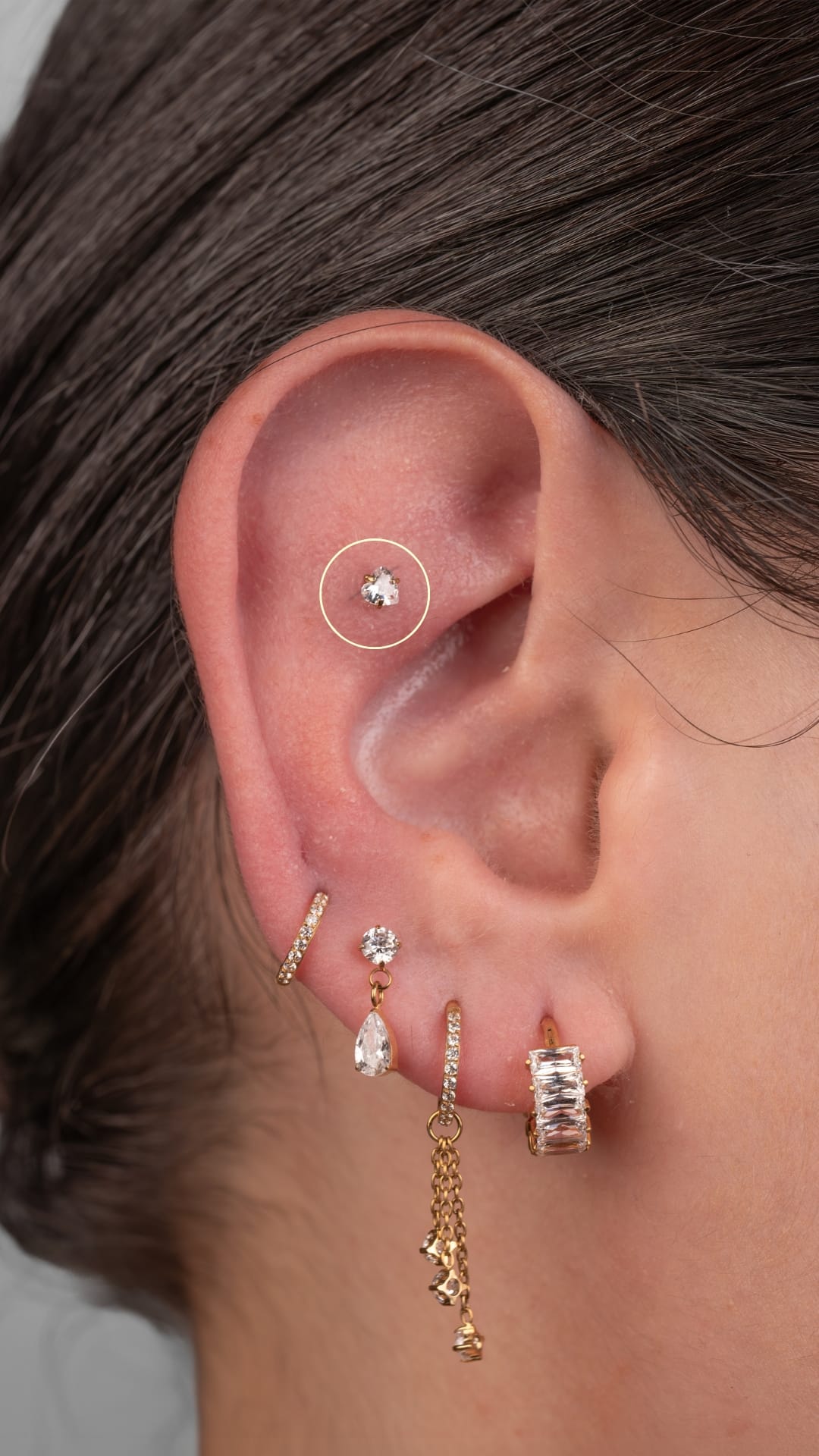 En un primer plano se aprecia una oreja decorada al estilo Noble Art, en la que destaca el pendiente «Flat Helix» de NOBLE ART OFFICIAL COORP entre pendientes de oro y diamantes, aros con colgantes y un pequeño pendiente en el cartílago interno, todo ello realzado con un sutil detalle circular.
