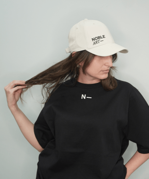 Una persona que lleva la camiseta negra «Noble Art by Noble Art», combinada con una gorra blanca de NOBLE ART, se aparta el pelo castaño hacia un lado y desvía la mirada, dejando entrever sutilmente un piercing sobre un fondo liso.