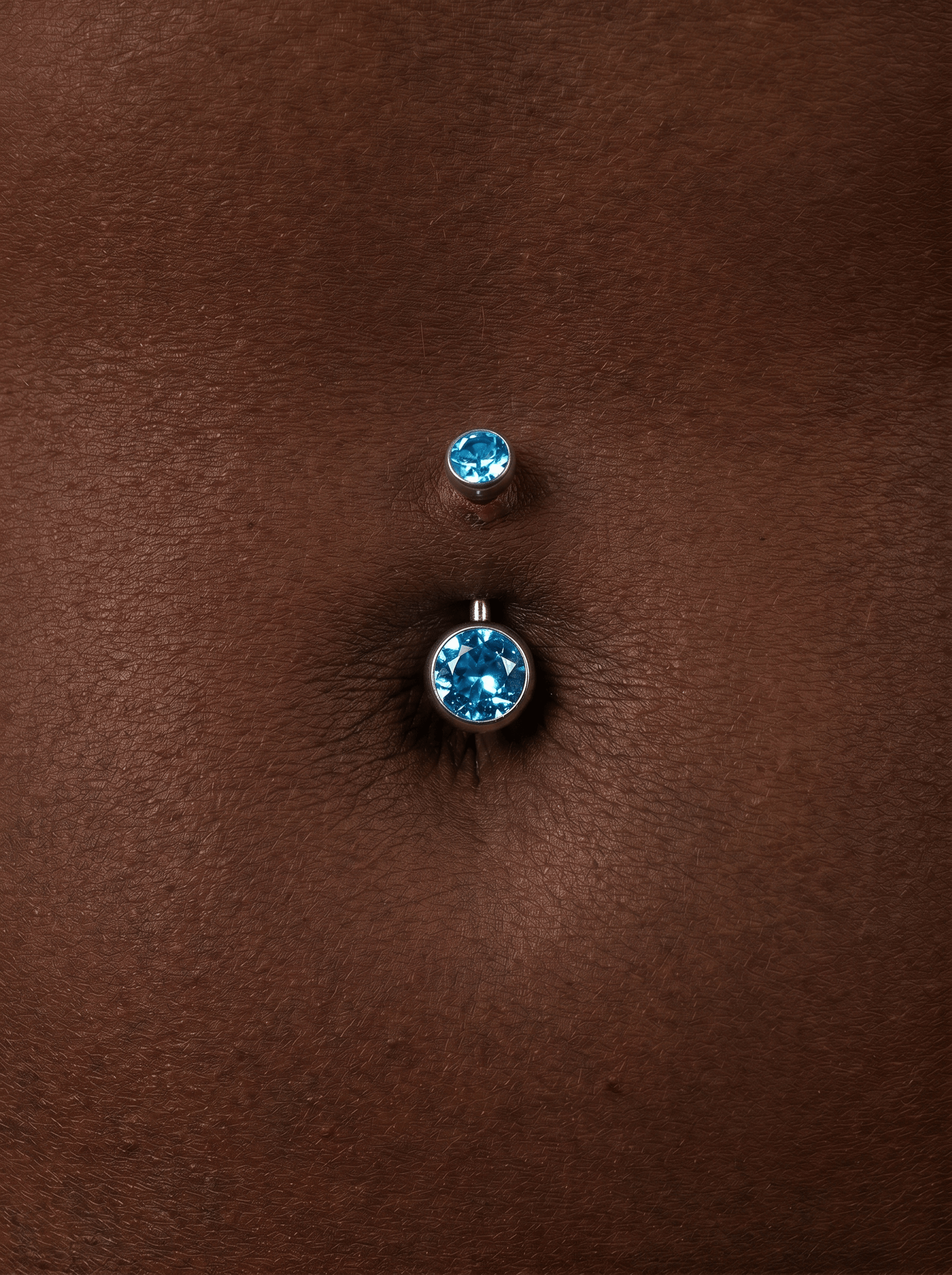 Eine Nahaufnahme des Bauchnabelpiercings „Ombligo cz azul“ von Titanio-Noble Art mit zwei blauen Kristallen, die in eine silberfarbene Fassung eingefasst sind und auf dunkelbrauner Haut funkeln.