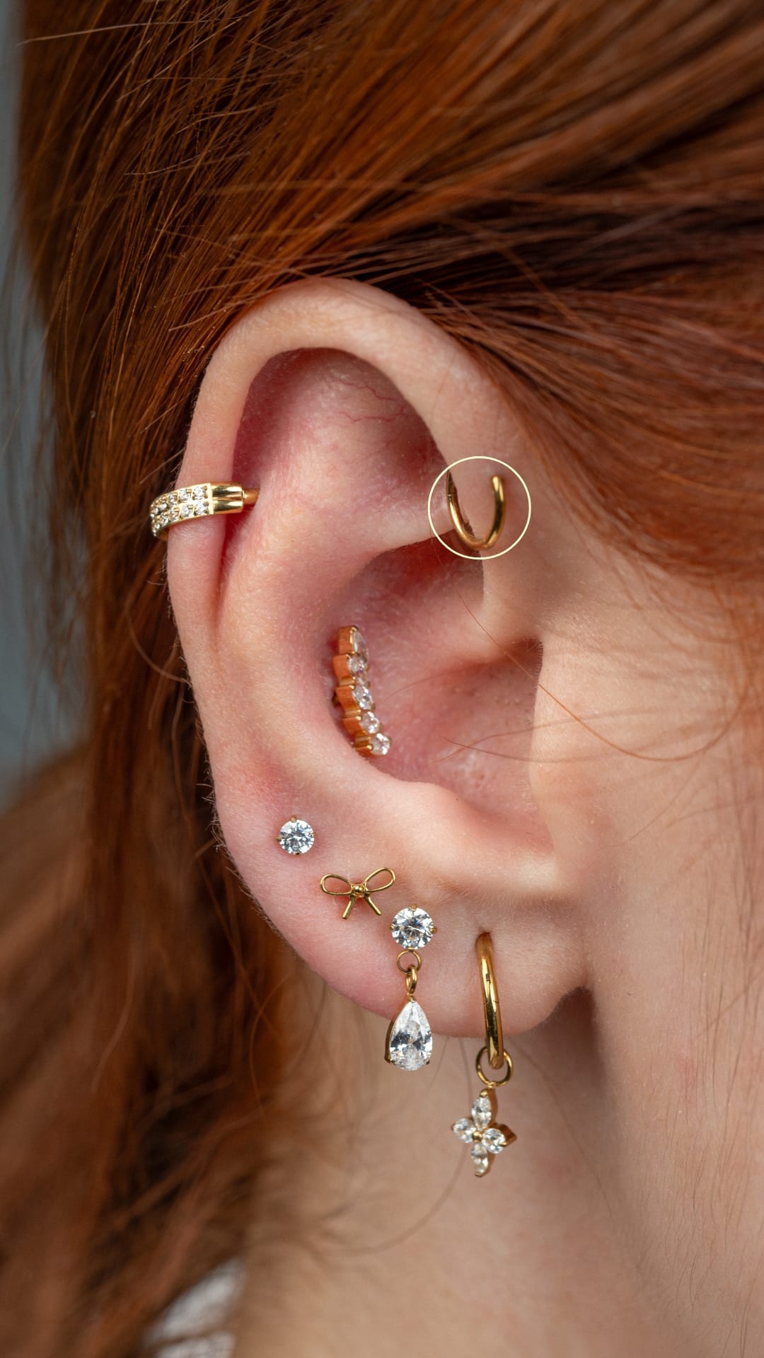Un primer plano de la oreja de una persona con múltiples piercings —en el que destaca el modelo «Fordward Helix» de NOBLE ART OFFICIAL COORP—: aros de oro y piedras preciosas, pendientes de botón y colgantes que resaltan sobre el cabello de color marrón rojizo, poniendo de relieve la refinada ornamentación de la oreja.