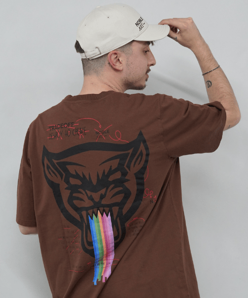 Un hombre con una gorra blanca, al que se ve de espaldas ajustándose la visera, lleva la «Camiseta Arcoíris» de Noble Art, una camiseta marrón con un gran dibujo de una pantera negra, una franja arcoíris que sale de su boca, diseños inspirados en tatuajes y texto en rojo.