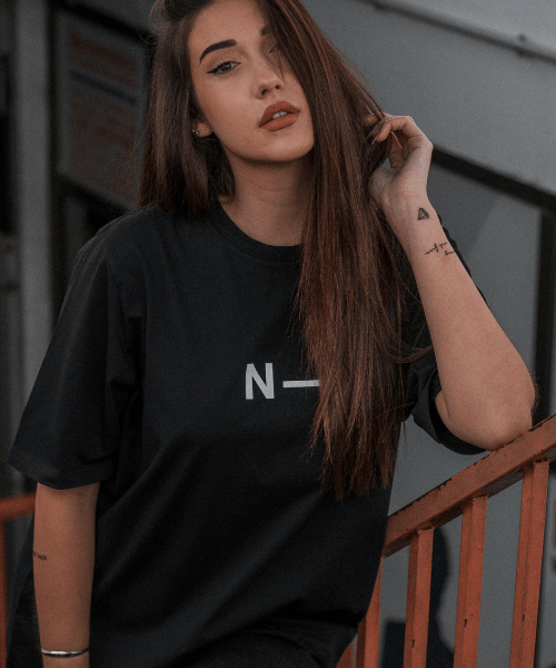 Una joven con el pelo largo y castaño y muy poco maquillaje posa sobre una barandilla naranja, vestida con una camiseta negra holgada de Noble Art by Noble Art, en la que se aprecian pequeños tatuajes en la muñeca izquierda y el antebrazo.