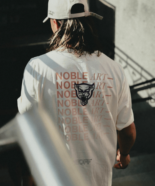 Una persona con el pelo largo, un tatuaje y una gorra blanca aparece de espaldas a la cámara con puesta la camiseta «Noble Art Camiseta Trade X Noble Art», en la que se repite el nombre de la marca y aparece el dibujo estilizado de un lobo bajo la luz del sol.
