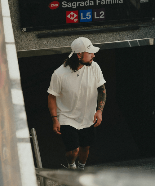 Un hombre que lleva una camiseta de Noble Art Camiseta Trade X, una gorra blanca y unos pantalones cortos negros sube las escaleras del metro en dirección a un estudio de piercings o tatuajes. Detrás de él, un cartel indica «Sagrada Familia» y señala las líneas de metro L5 y L2.