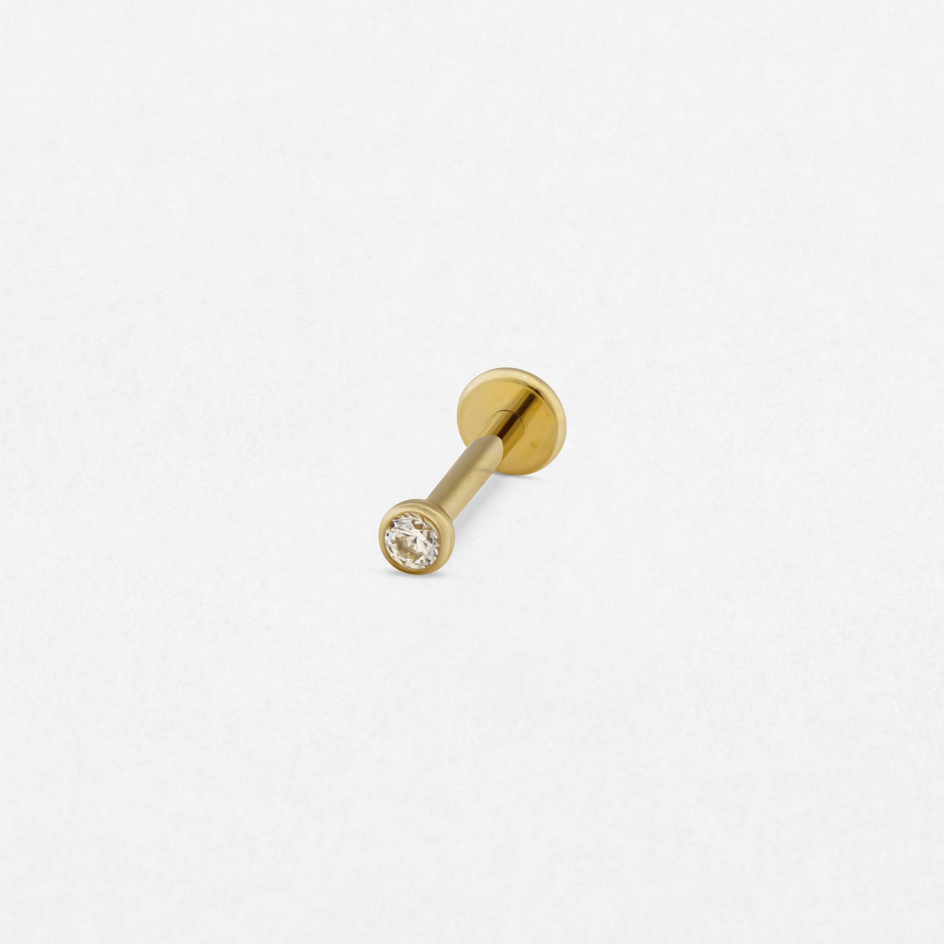 El pendiente «Circonita blanca / oro» de Titanio-Noble Art es un pequeño pendiente de botón con la parte trasera plana, adornado con una única piedra preciosa redonda y transparente, que destaca sobre un fondo blanco liso.