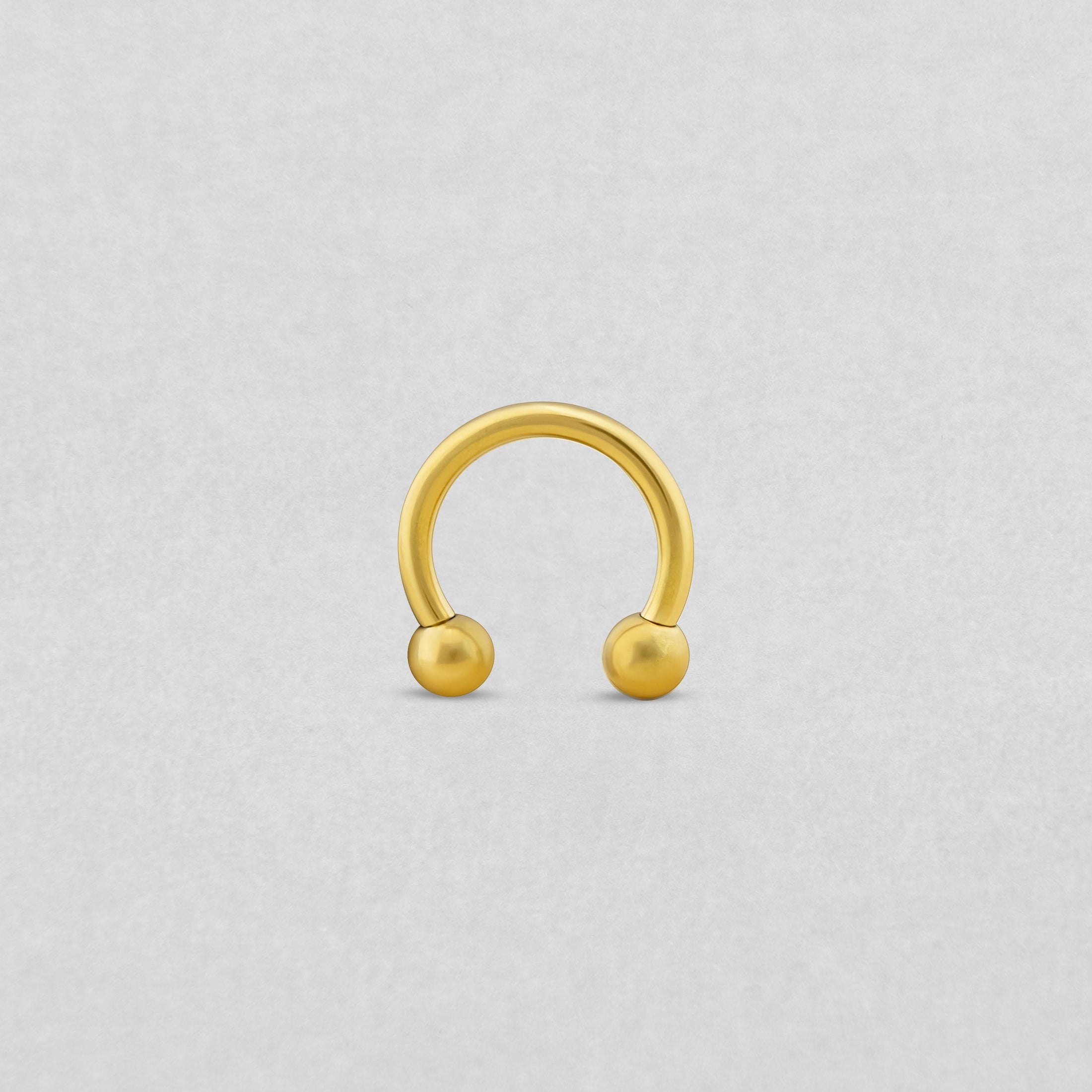 La barra Circular de NOBLE ART BEAUTY SOCIEDAD LIMITADA presenta un acabado dorado con cuentas redondas en cada extremo, y se muestra en posición vertical sobre un fondo gris claro.