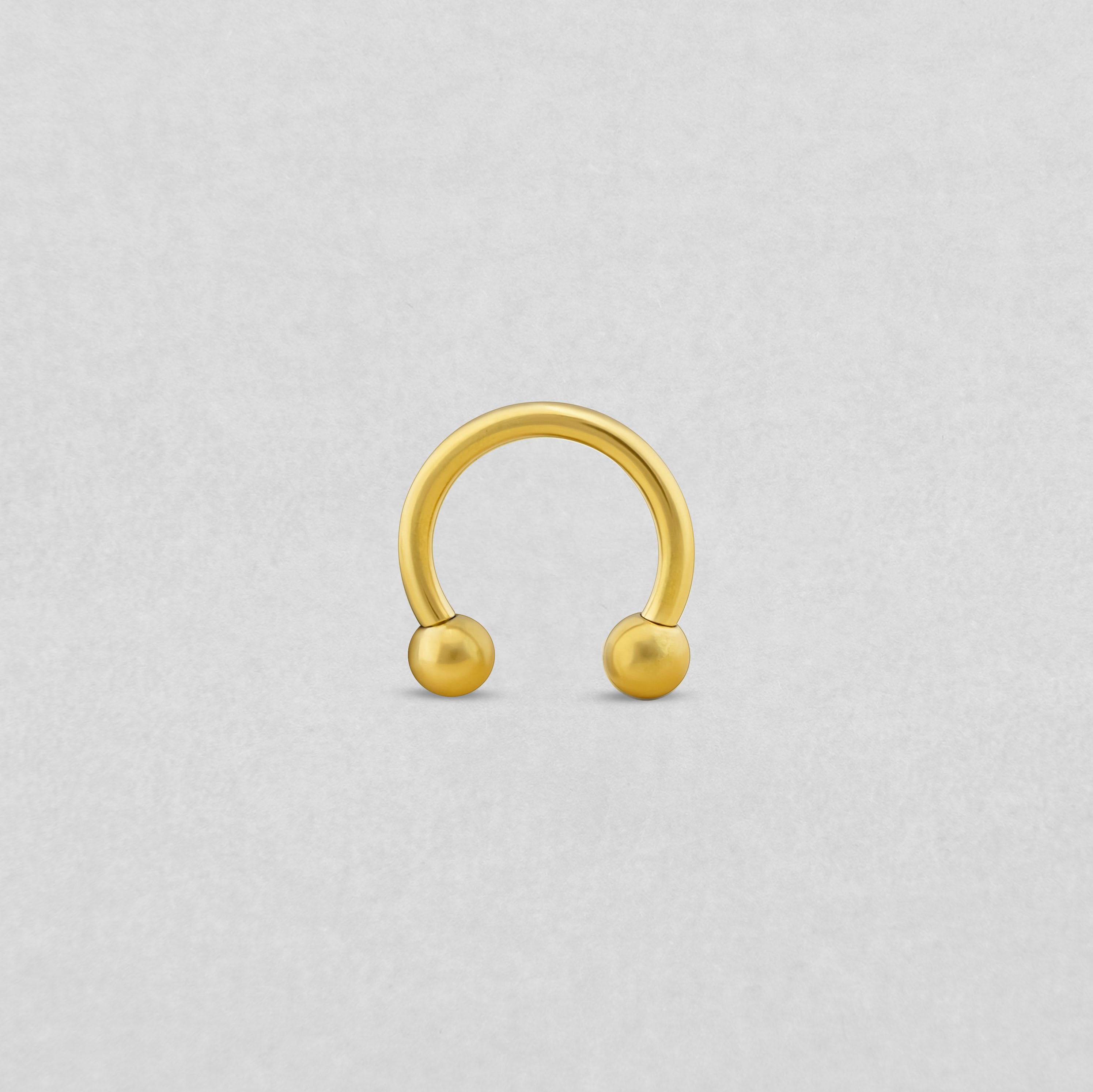 La barra Circular de NOBLE ART BEAUTY SOCIEDAD LIMITADA presenta un acabado dorado con cuentas redondas en cada extremo, y se muestra en posición vertical sobre un fondo gris claro.