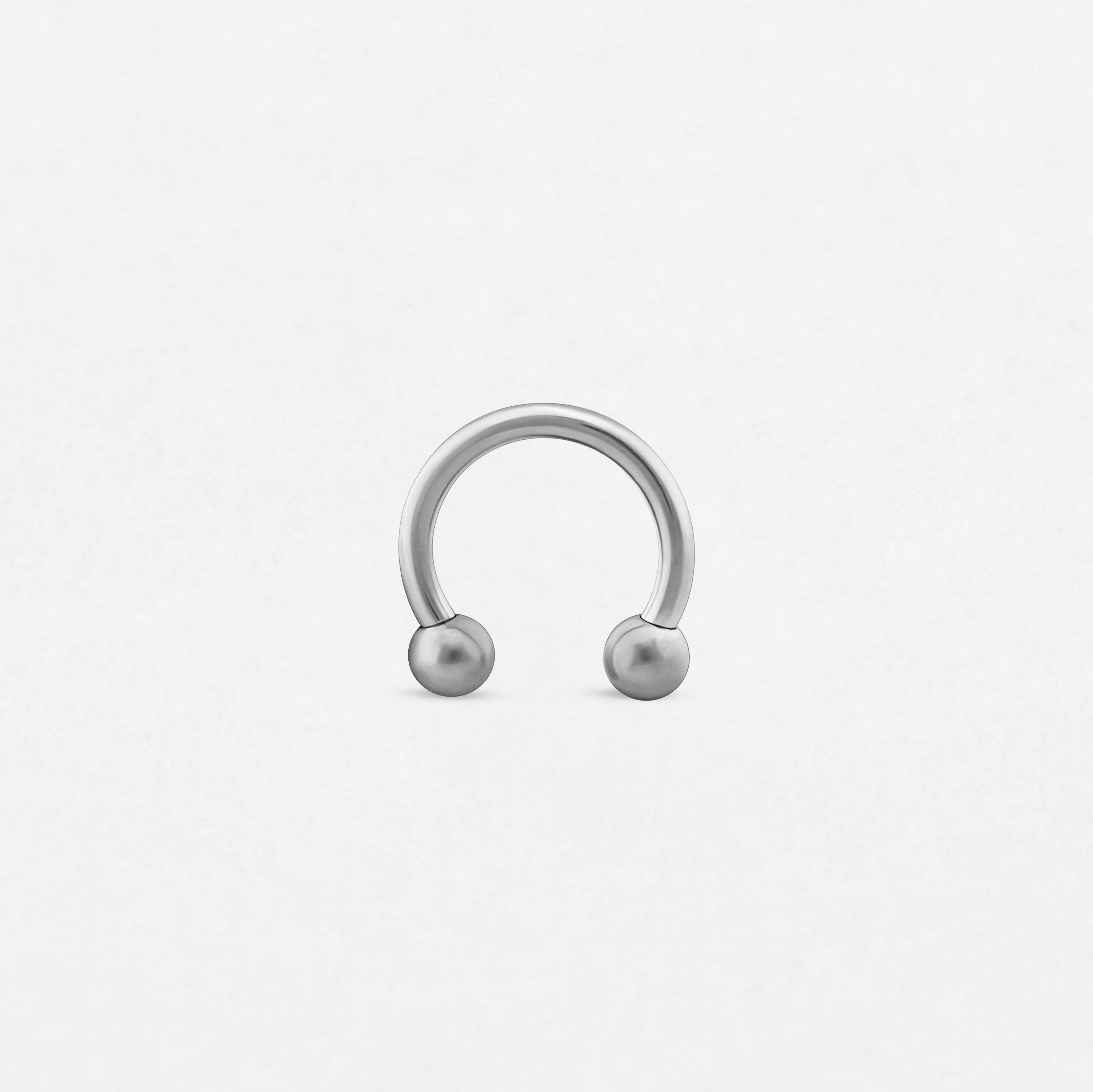 Se muestra, en posición vertical sobre un fondo blanco liso, una barra circular «Titanio-Noble Art» de plata, con forma de herradura y una bola en cada extremo.
