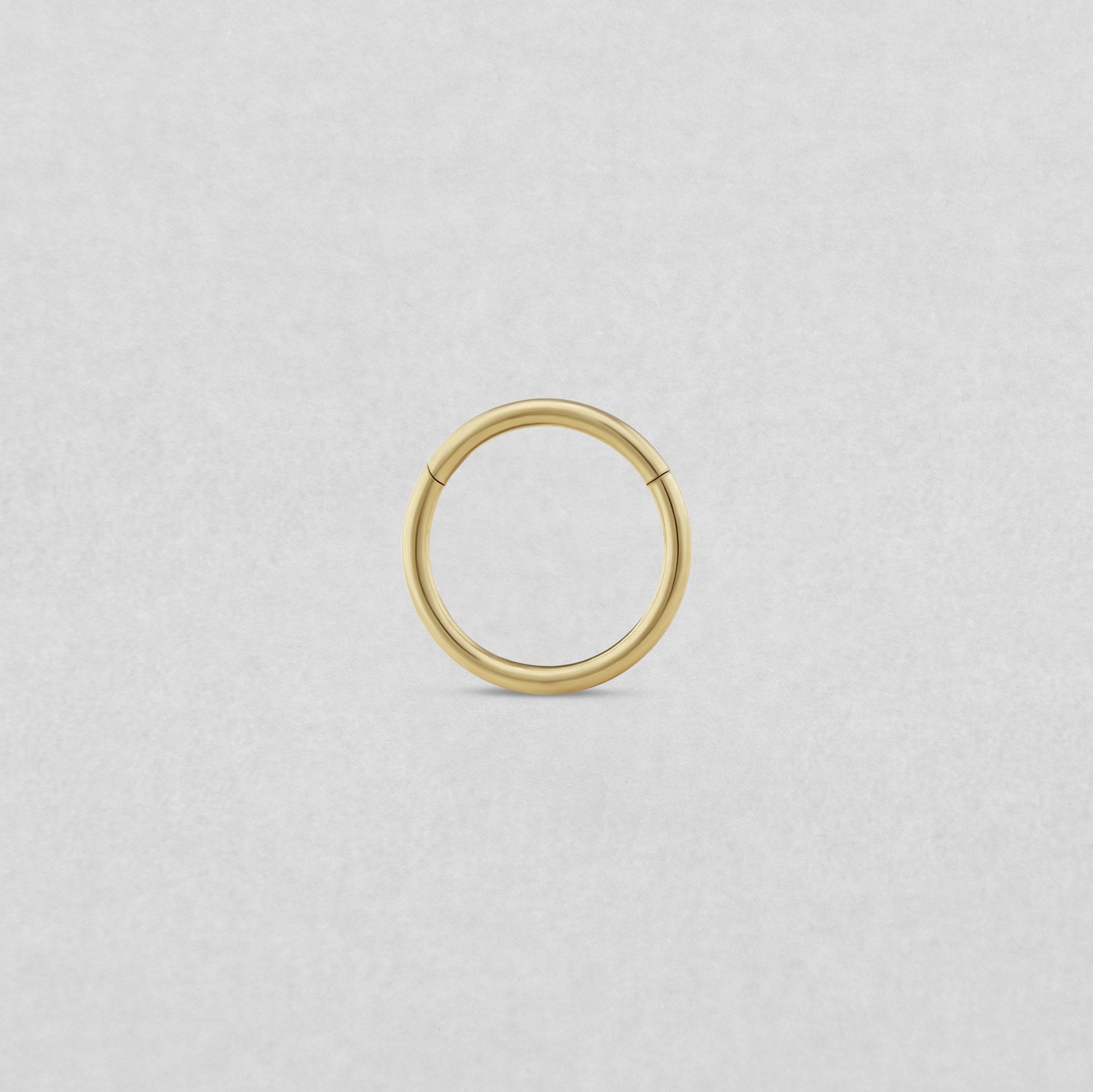 El «Clicker básico» de NOBLE ART BEAUTY SOCIEDAD LIMITADA es un pequeño pendiente de aro dorado sin costuras para perforaciones, presentado sobre un fondo liso de color gris claro.