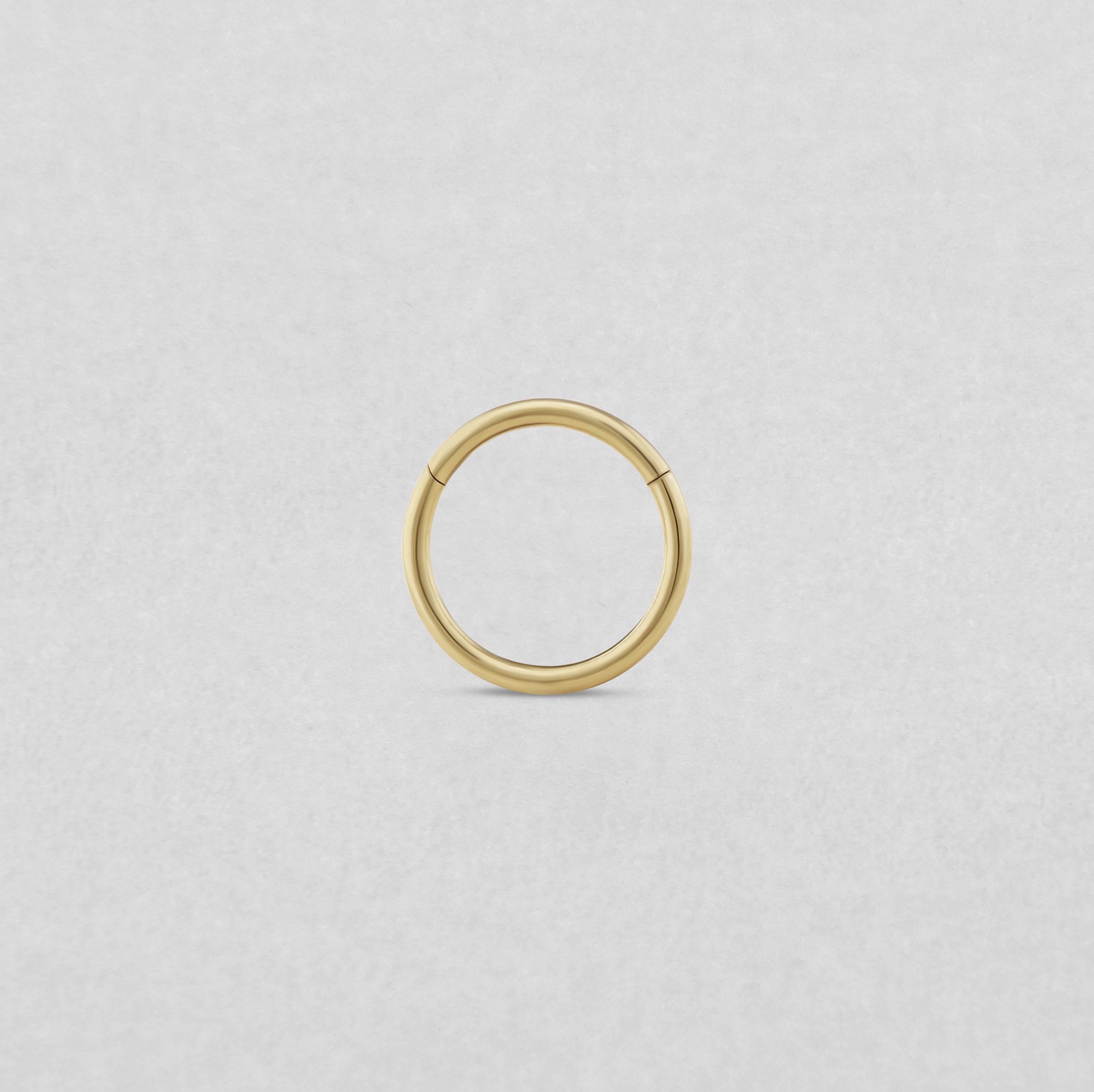 El «Clicker básico» de NOBLE ART BEAUTY SOCIEDAD LIMITADA es un pequeño pendiente de aro dorado sin costuras para perforaciones, presentado sobre un fondo liso de color gris claro.