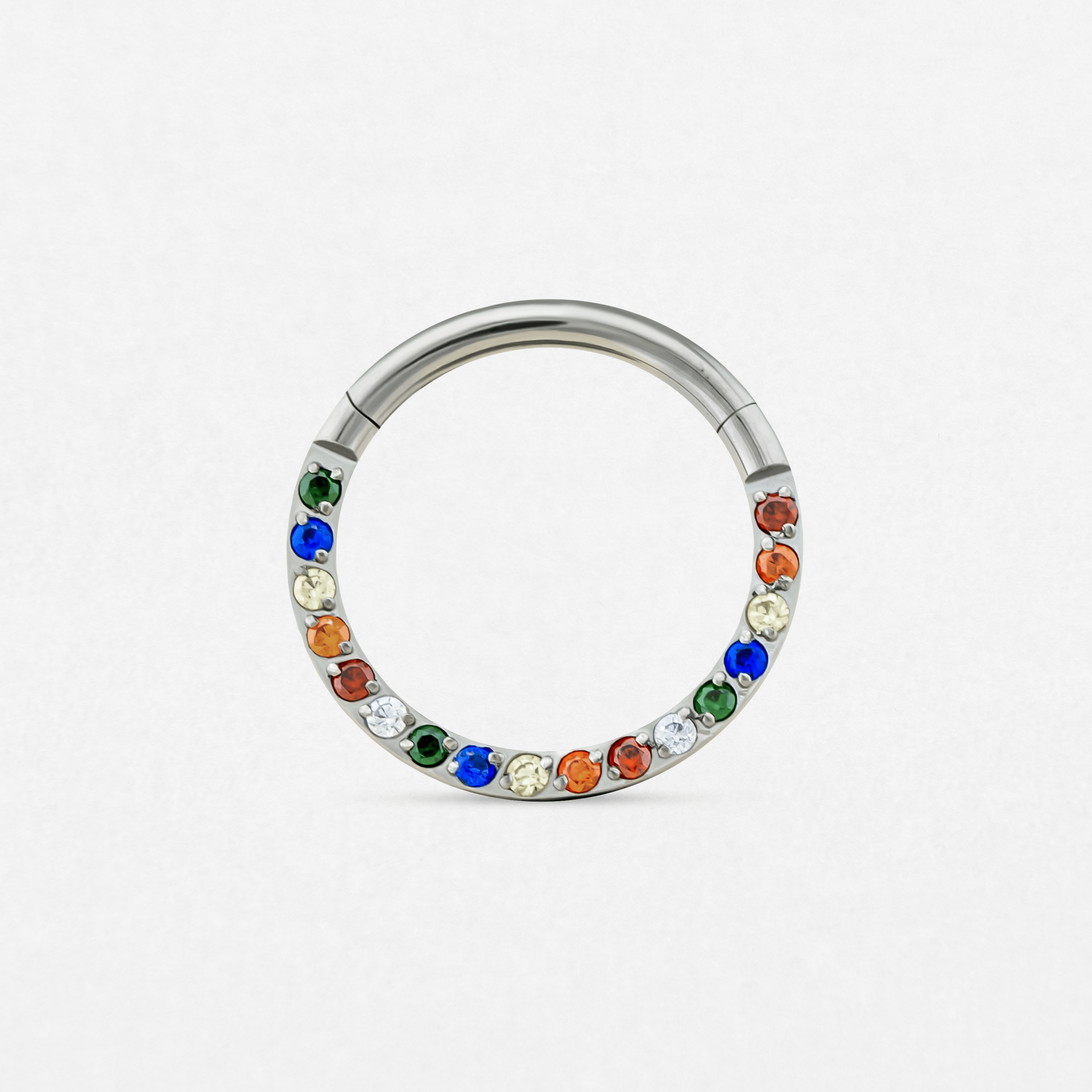 El Clicker arcoiris de Titanio-Noble Art es un anillo circular de plata para piercing con gemas azules, verdes, rojas y transparentes dispuestas alternativamente a lo largo de la mitad de la banda. Un accesorio elegante sobre fondo blanco, ideal para combinar con otras joyas o tras la eliminación de tatuajes con láser.