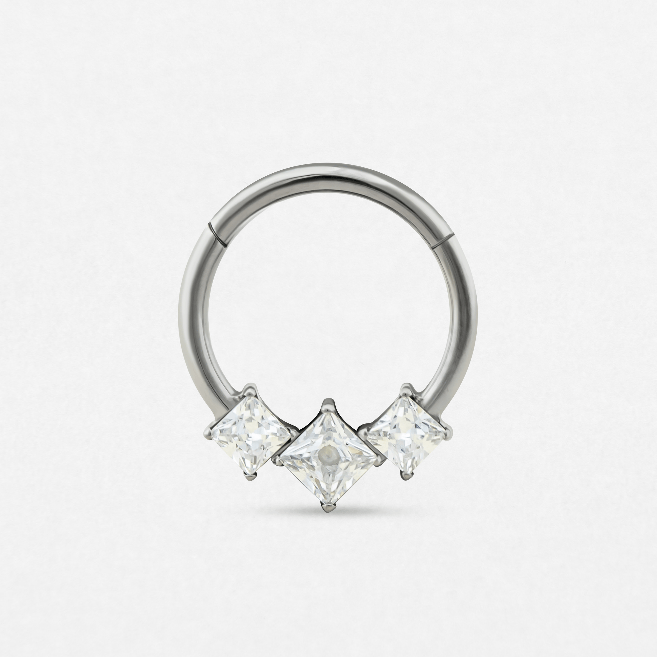 El anillo «Clicker Eros» de Titanio-Noble Art es un anillo circular de plata con tres piedras preciosas transparentes de talla cuadrada, distribuidas a intervalos regulares, que refleja la elegancia de las joyas modernas para piercings, representadas sobre un fondo blanco liso.
