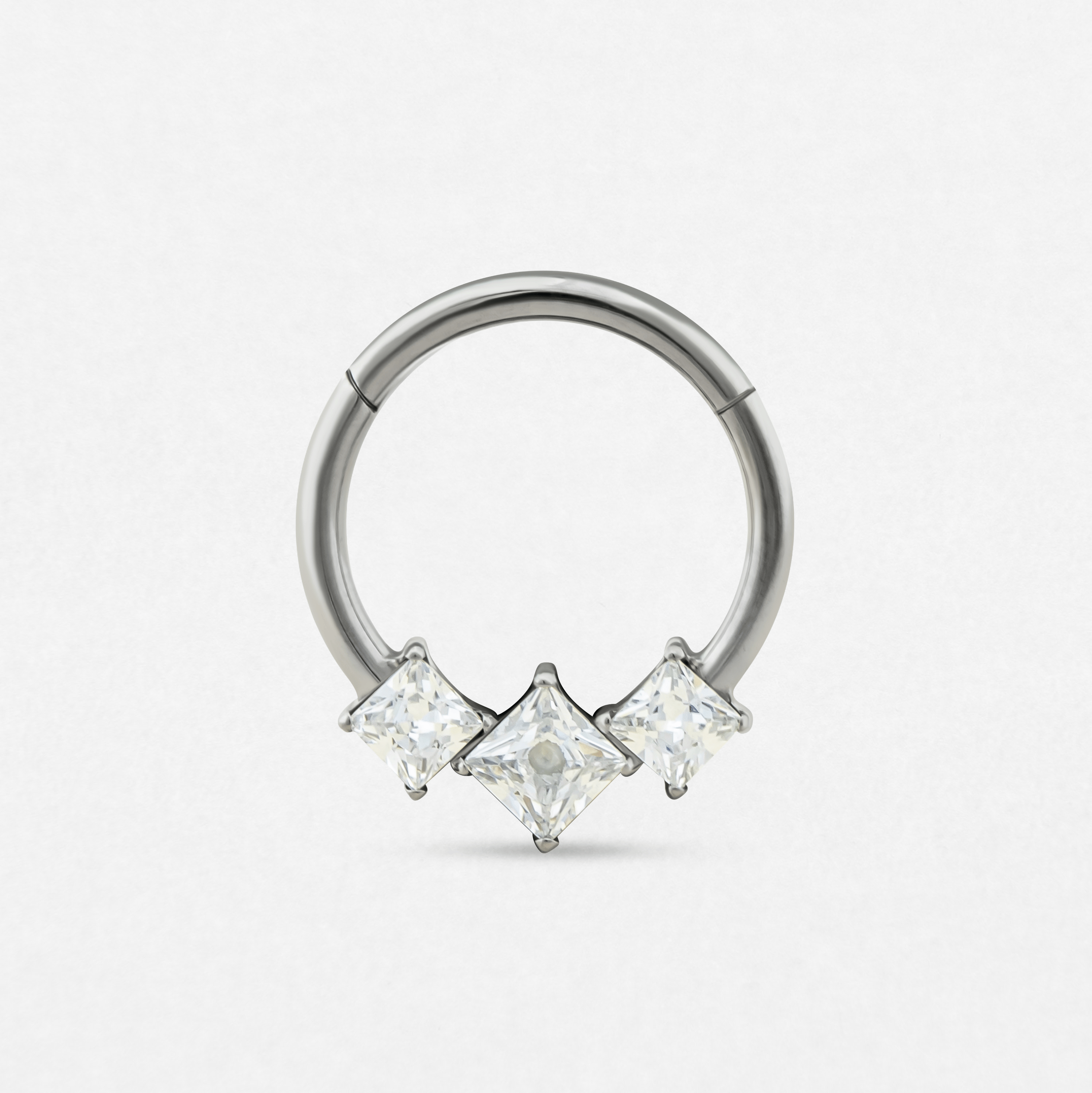 El anillo «Clicker Eros» de Titanio-Noble Art es un anillo circular de plata con tres piedras preciosas transparentes de talla cuadrada, distribuidas a intervalos regulares, que refleja la elegancia de las joyas modernas para piercings, representadas sobre un fondo blanco liso.