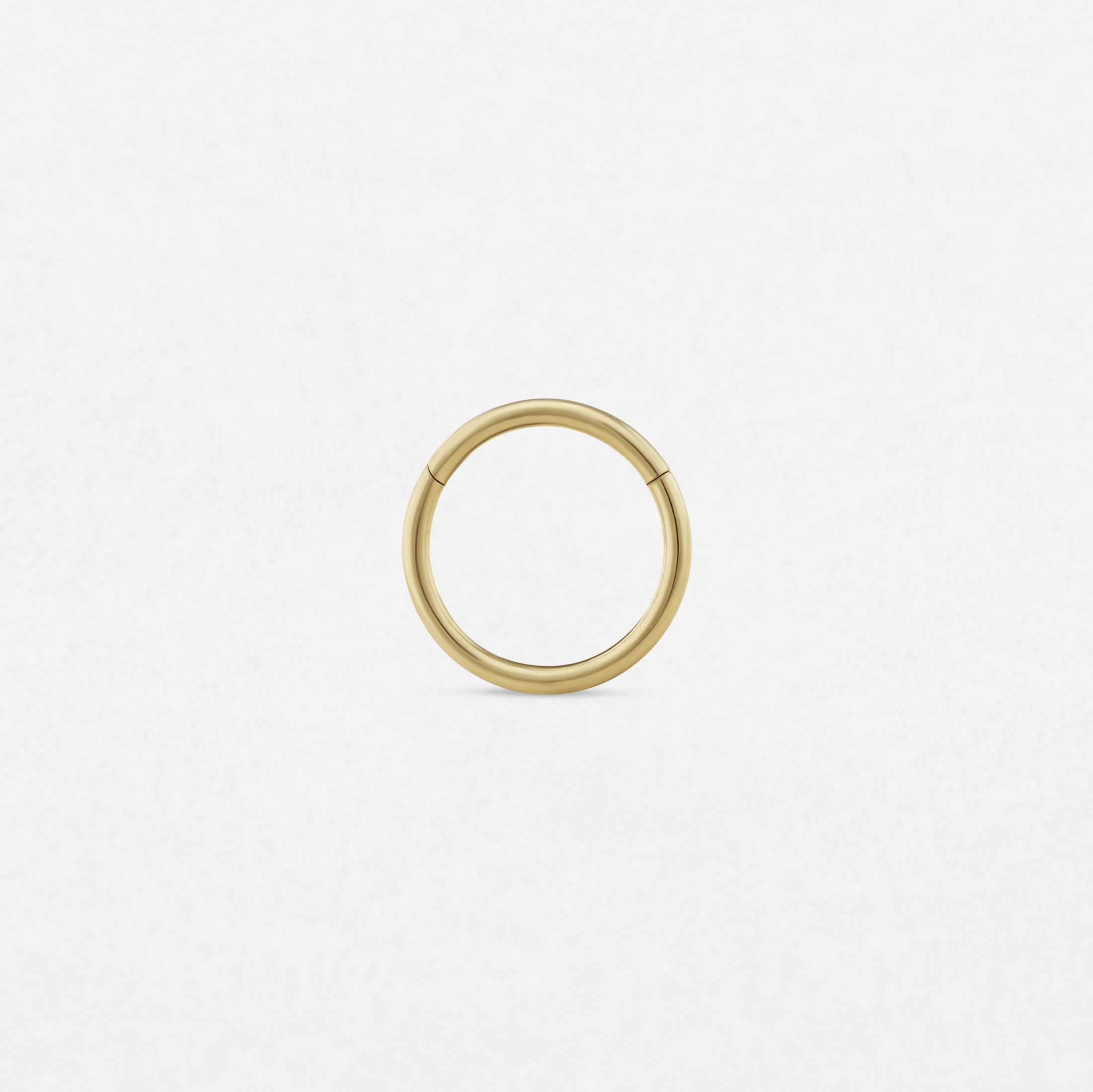 Un aro «Clicker» minimalista y sin costuras, de color dorado, de la marca Titanio-Noble Art, destaca sobre un fondo blanco liso, lo que le confiere un estilo refinado gracias a su sencillo diseño circular.