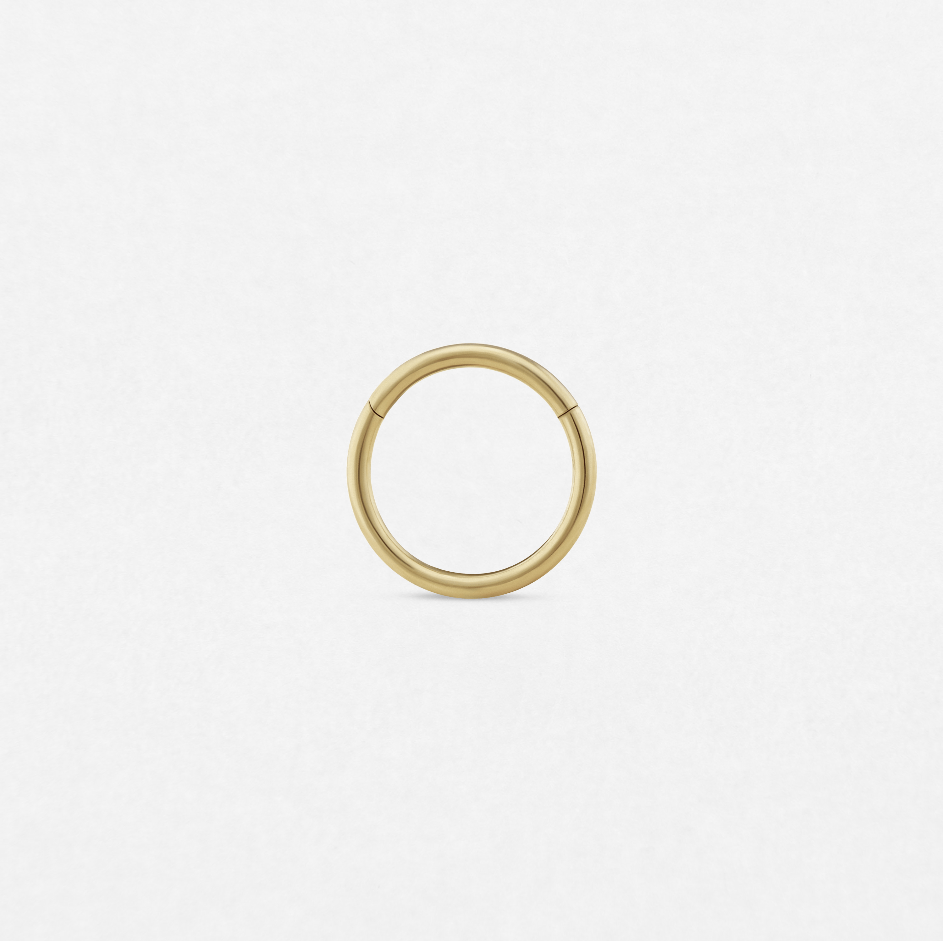 Un aro «Clicker» minimalista y sin costuras, de color dorado, de la marca Titanio-Noble Art, destaca sobre un fondo blanco liso, lo que le confiere un estilo refinado gracias a su sencillo diseño circular.