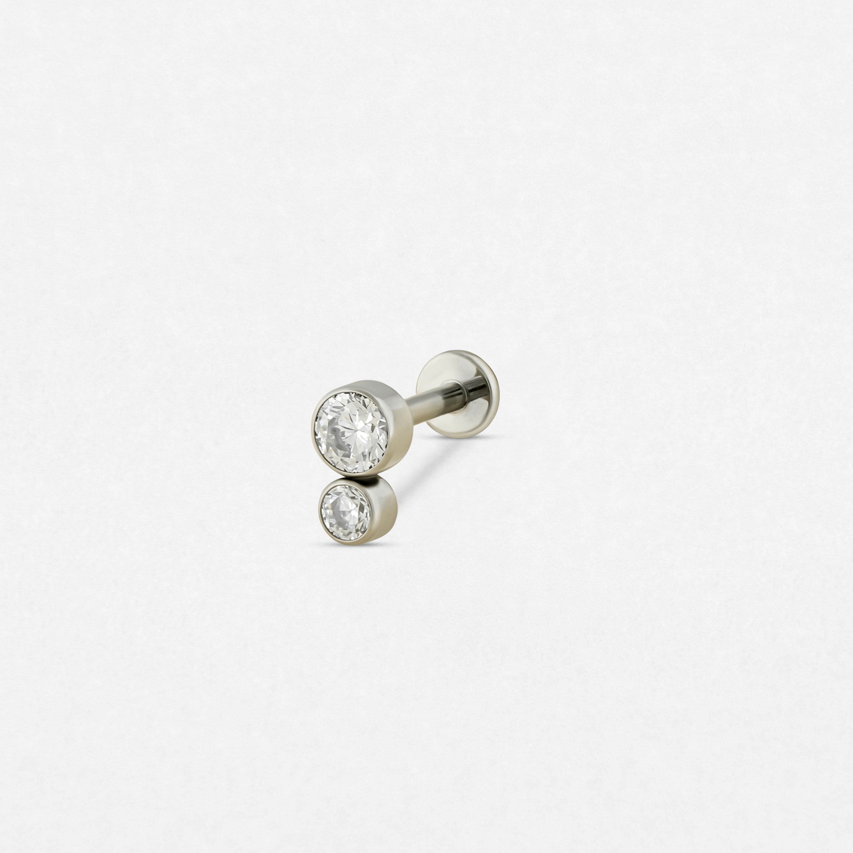 El modelo «Cluster blanco 2cz» de Titanio-Noble Art es un pendiente de botón de plata con dos piedras preciosas redondas y transparentes de diferentes tamaños engastadas en bisel, dispuestas elegantemente en vertical sobre un fondo blanco.