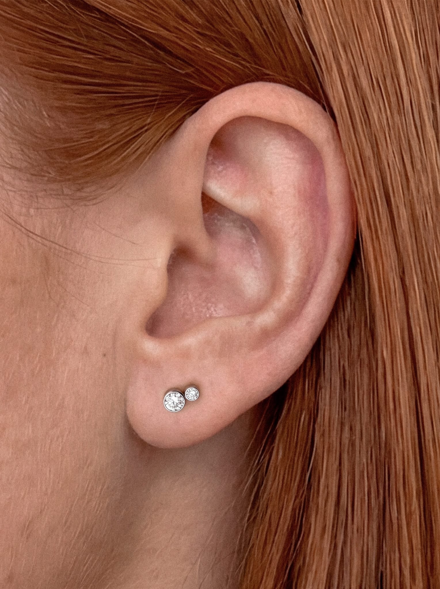 Un primer plano de una oreja adornada con el modelo «Cluster blanco 2cz» de Titanio-Noble Art: dos pequeños pendientes de botón redondos de plata con circonitas cúbicas transparentes. El elegante diseño resalta el piercing, que combina a la perfección con el pelo liso de color castaño rojizo de la persona.
