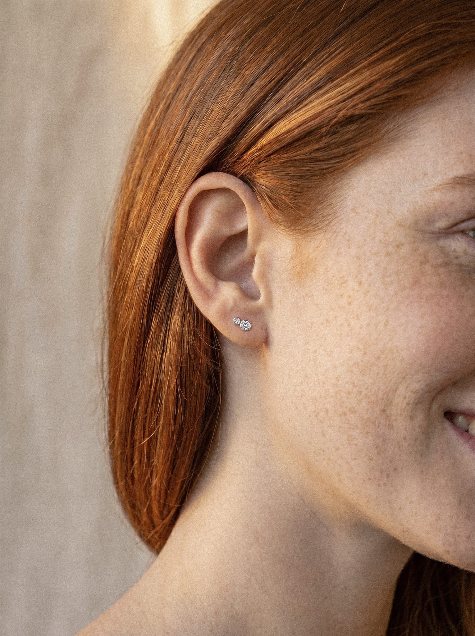 Primer plano de una persona con el pelo largo y pelirrojo y pecas claras, sonriendo. Lleva puestos los pendientes pequeños «Cluster blanco 2cz» de Titanio-Noble Art, con forma de flores y elaborados con circonitas cúbicas para aportar mayor brillo. El fondo, ligeramente desenfocado, centra la atención en la oreja y en el elegante diseño de los pendientes.