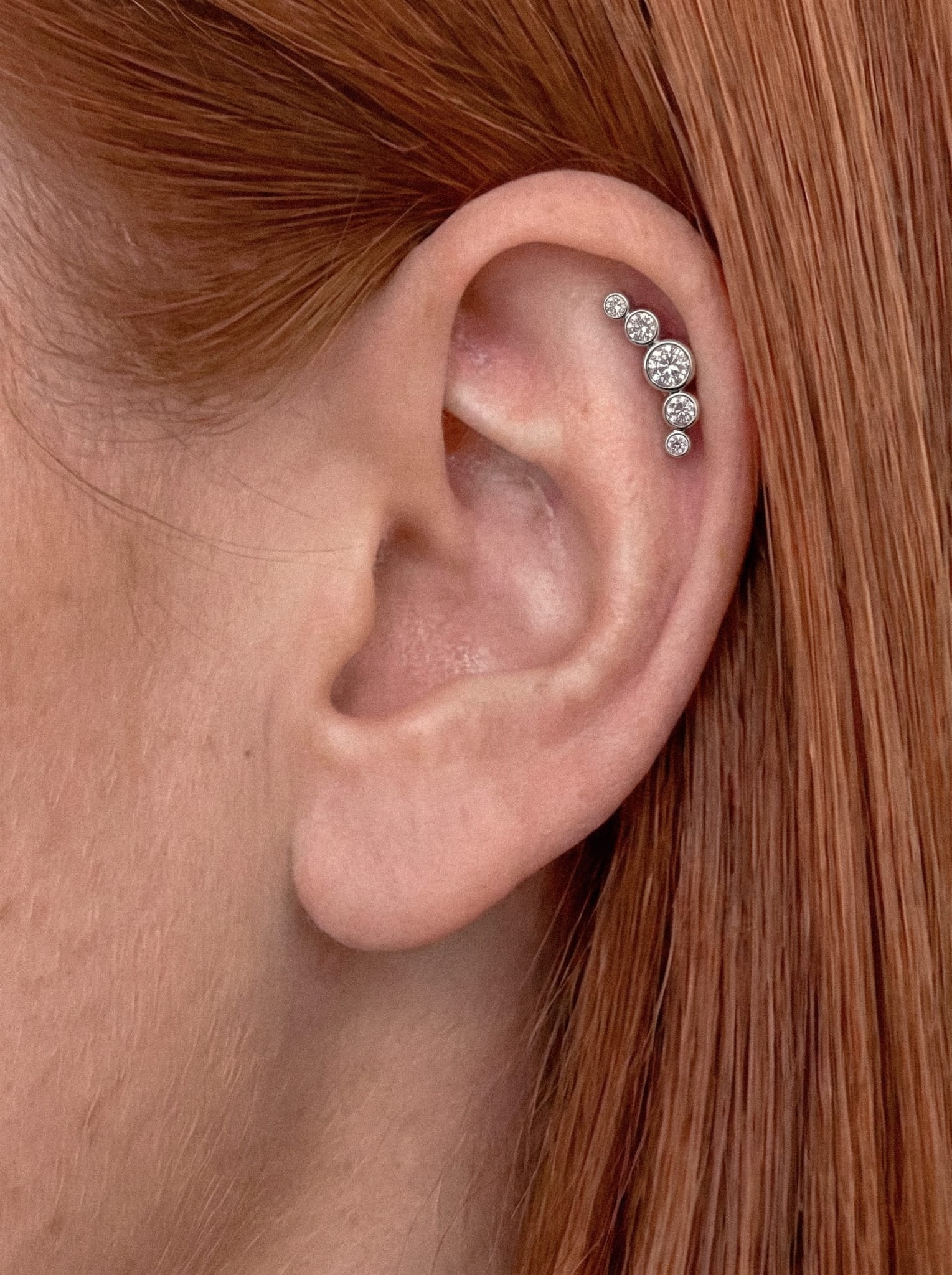 Un primer plano de la oreja de una persona adornada con el modelo «Ornamental 3 cz» de Titanio-Noble Art, en el que se aprecian unos brillantes pendientes de circonitas redondas dispuestos en una elegante curva a lo largo del cartílago, incluyendo un llamativo piercing con tres cristales. El pelo liso, de color castaño rojizo, enmarca la oreja, aportando un toque galáctico a este llamativo look metálico.