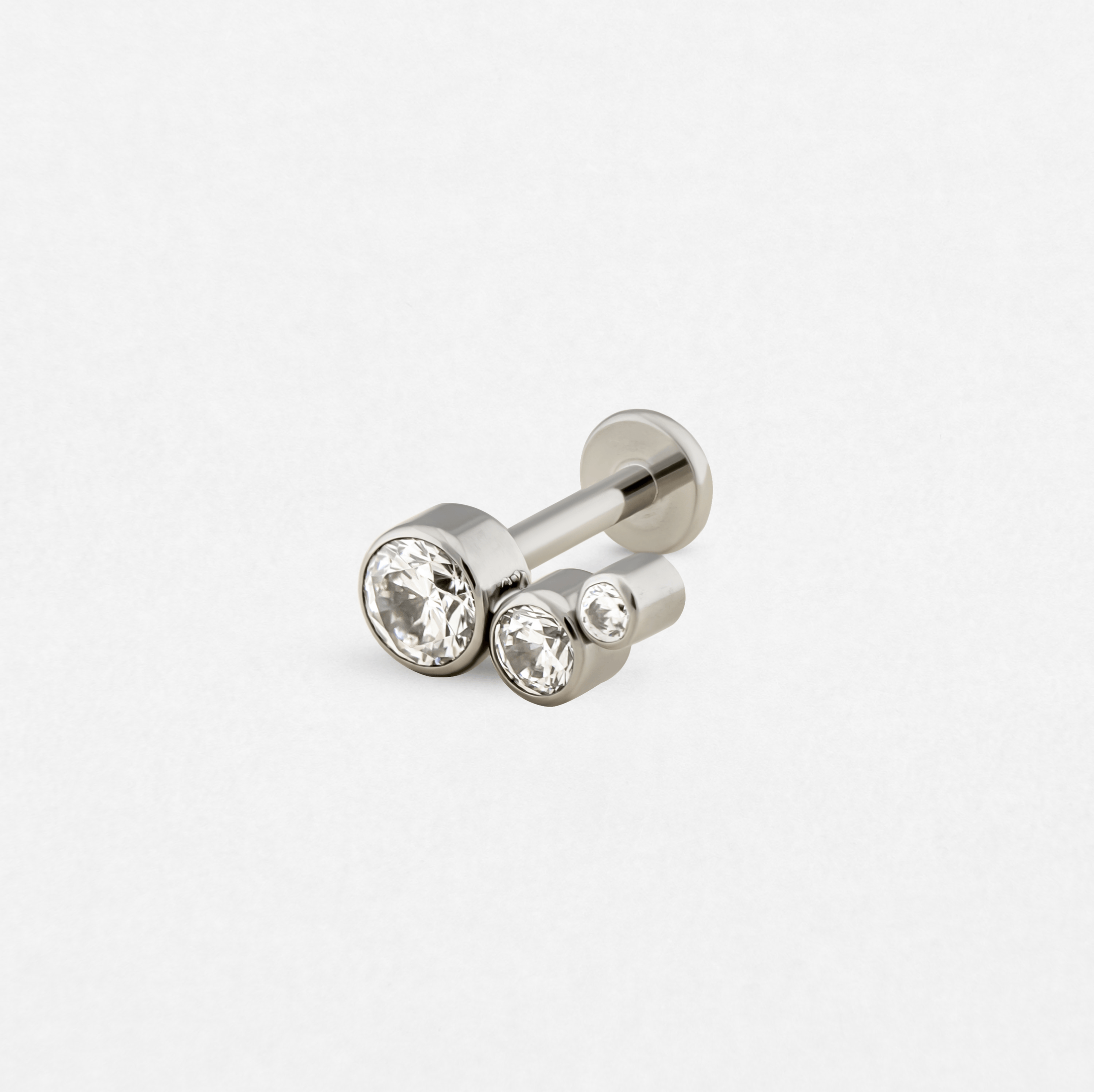 El pendiente de botón «Titanio- Noble Art Cluster decreciente» es un elegante pendiente de plata con tres piedras preciosas redondas y transparentes de diferentes tamaños dispuestas en fila, engastadas en un diseño metálico minimalista sobre un fondo blanco liso.