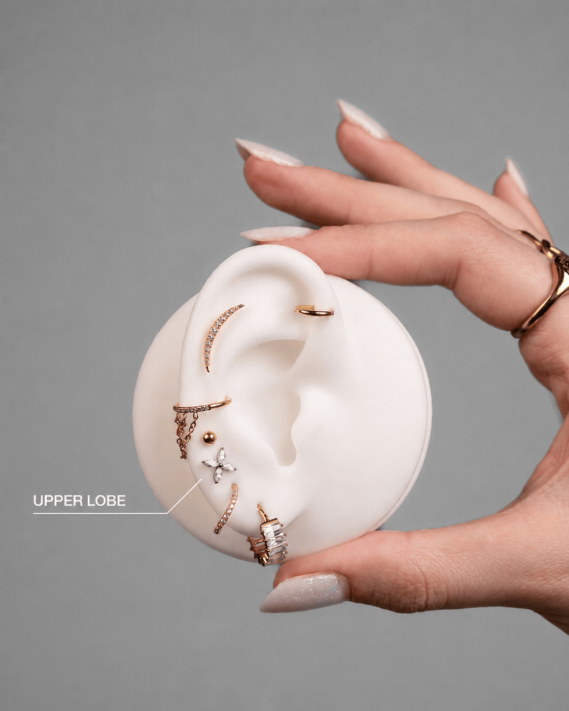 Una mano bien cuidada sostiene un modelo de oreja blanco adornado con los pendientes dorados y plateados UPPER LOBE de NOBLE ART OFFICIAL COORP, con una etiqueta que destaca la zona del lóbulo superior al estilo característico de Noble Art.