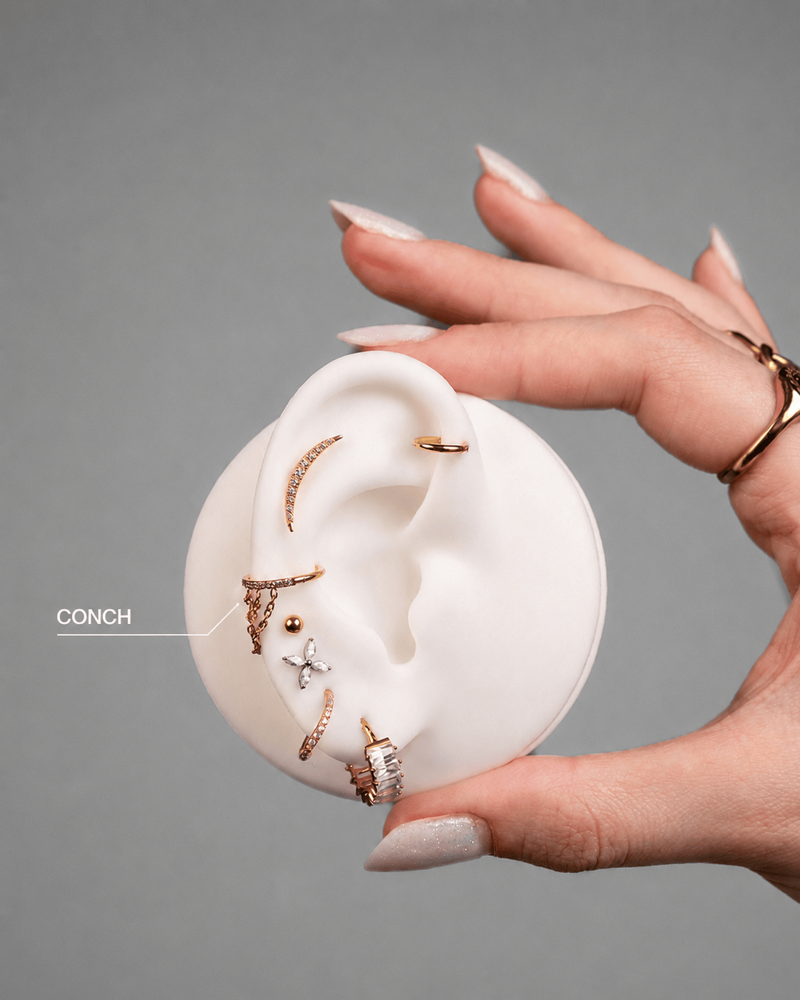 A well-groomed hand holds a white ear model with «CONCH» earrings by NOBLE ART OFFICIAL COORP in gold and diamonds. A label indicates the conch piercing, suitable for those considering laser removal after getting a tattoo. Plain grey background.