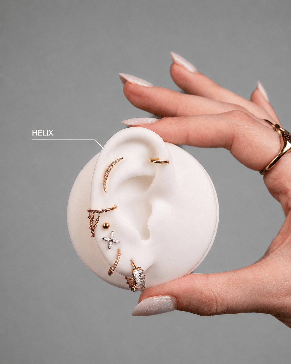 A well-groomed hand displays a white ear model with gold HELIX earrings by NOBLE ART OFFICIAL COORP, highlighting the upper helix piercing: the perfect inspiration for your next NOBLE ART OFFICIAL COORP HELIX piercing or tattoo.