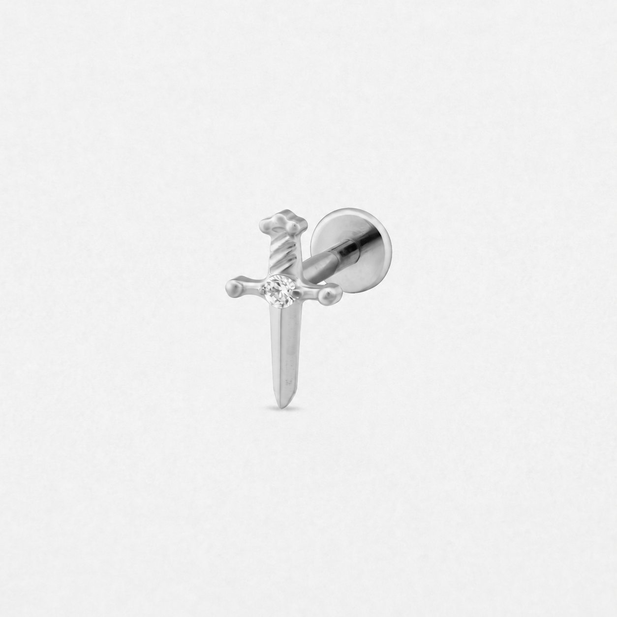 El modelo «Daga» de Titanio-Noble Art es un pendiente de botón plateado con forma de espada, con piedras preciosas transparentes en la hoja y la empuñadura, perfecto para los amantes de los piercings o los seguidores de Noble Art. Se muestra sobre un fondo blanco liso.