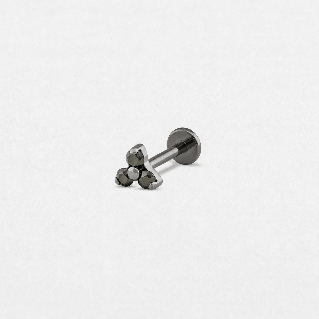 El modelo «Trinity cz» de Titanio-Noble Art es un pequeño pendiente de botón metálico con un cierre circular plano y una parte delantera en la que destaca un conjunto triangular formado por tres piedras preciosas negras, ideal para cualquier tipo de piercing, que se muestra sobre un fondo blanco liso.
