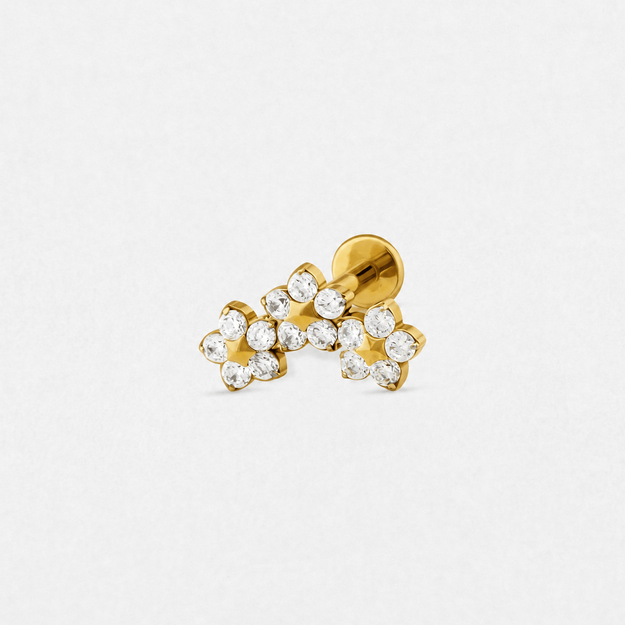 El pendiente «Triple Flower» de Titanio-Noble Art es un pendiente dorado con tres pequeñas formas de flor, cada una engastada con piedras preciosas redondas transparentes en un diseño curvo —perfecto para combinar con tu piercing favorito— y presentado sobre un fondo blanco.