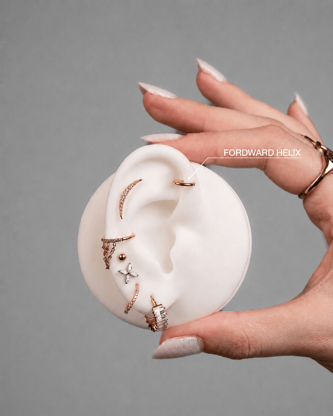 Una mano bien cuidada luce un modelo de oreja blanco adornado con piercings dorados de NOBLE ART OFFICIAL COORP; una etiqueta destaca la zona del «FORWARD HÉLIX», muy popular tras la eliminación con láser de un antiguo tatuaje o de un piercing anterior.