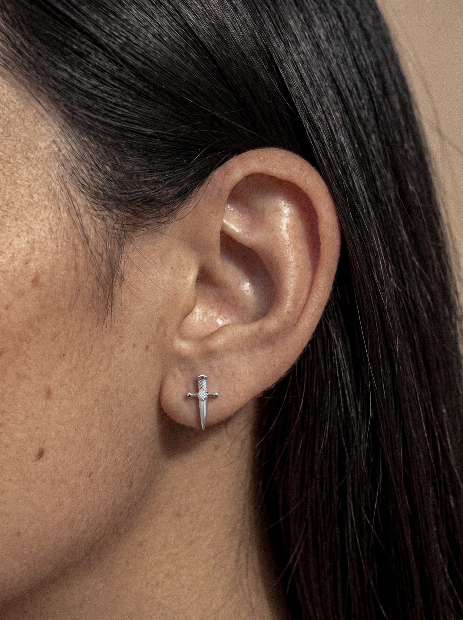 Primer plano de la oreja de una persona que lleva el pendiente de plata con forma de espada «Daga», de Titanio-Noble Art, un accesorio que evoca los «accesorios con carácter». El pelo oscuro de la persona está recogido detrás de la oreja, con un fondo neutro.