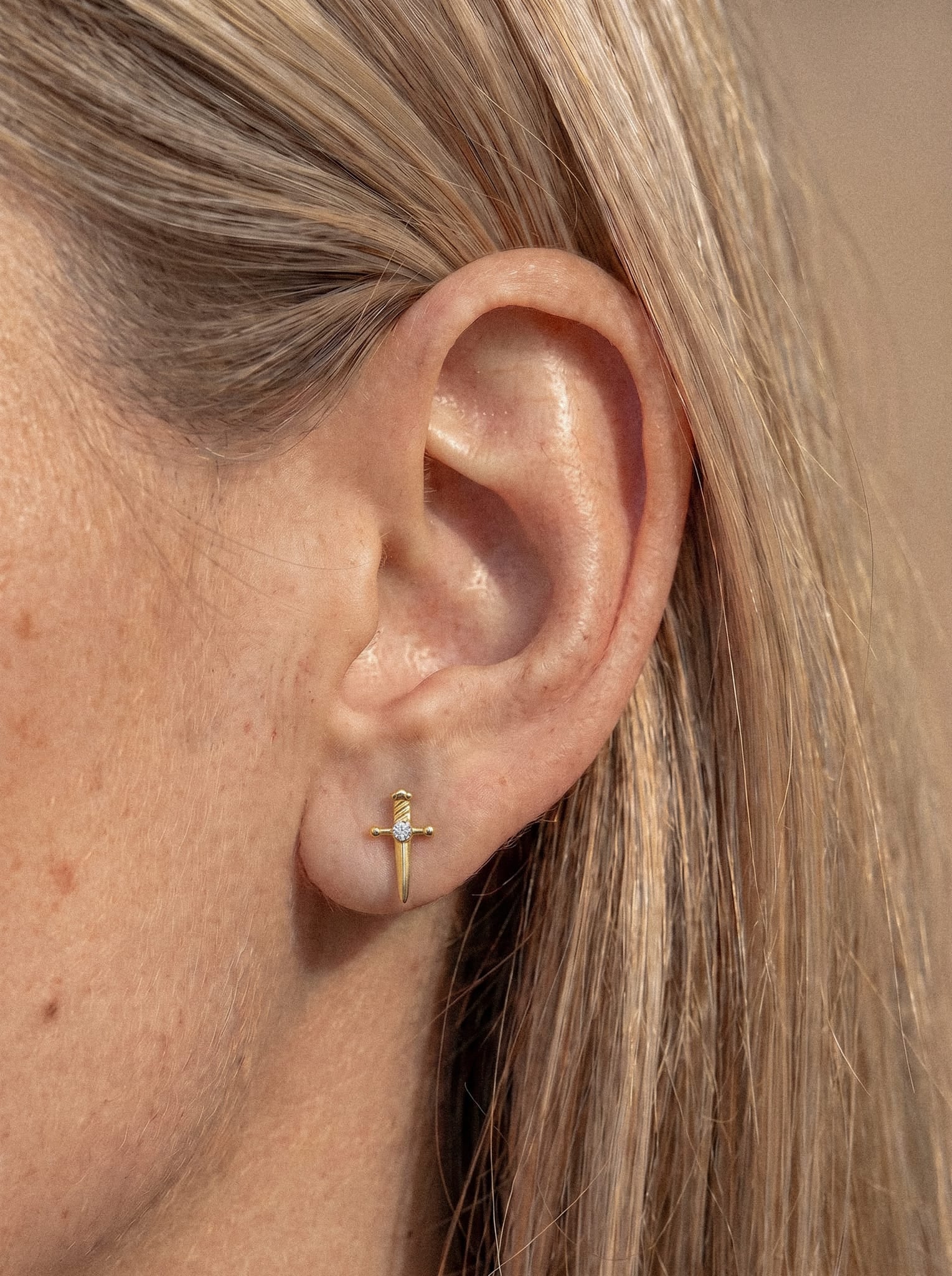 Un primer plano de la oreja de una persona con el pelo rubio y liso muestra cómo lleva puesto el pendiente de botón «Daga» de Titanio-Noble Art: un pequeño accesorio de oro con forma de cruz que recuerda a una mini daga dorada, con una delicada circonita en el centro que le aporta un sutil brillo.