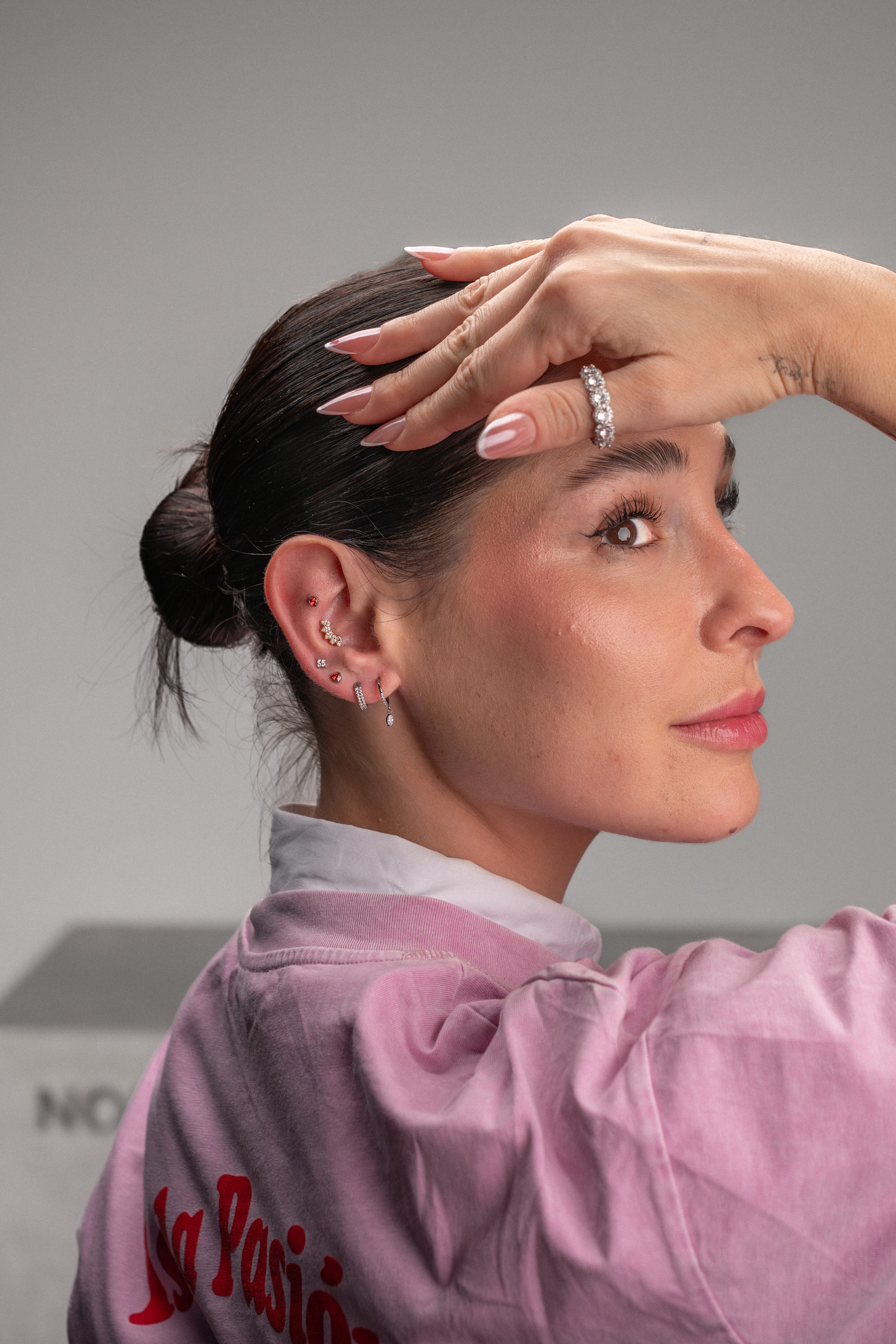 Una mujer de pelo oscuro, recogido en un moño bajo, posa de perfil, mostrando sus anillos y sus múltiples pendientes de agujero. Lleva una camiseta rosa con texto en rojo. Producto destacado: Circonita Rosa de Titanio-Noble Art. El fondo es gris liso.