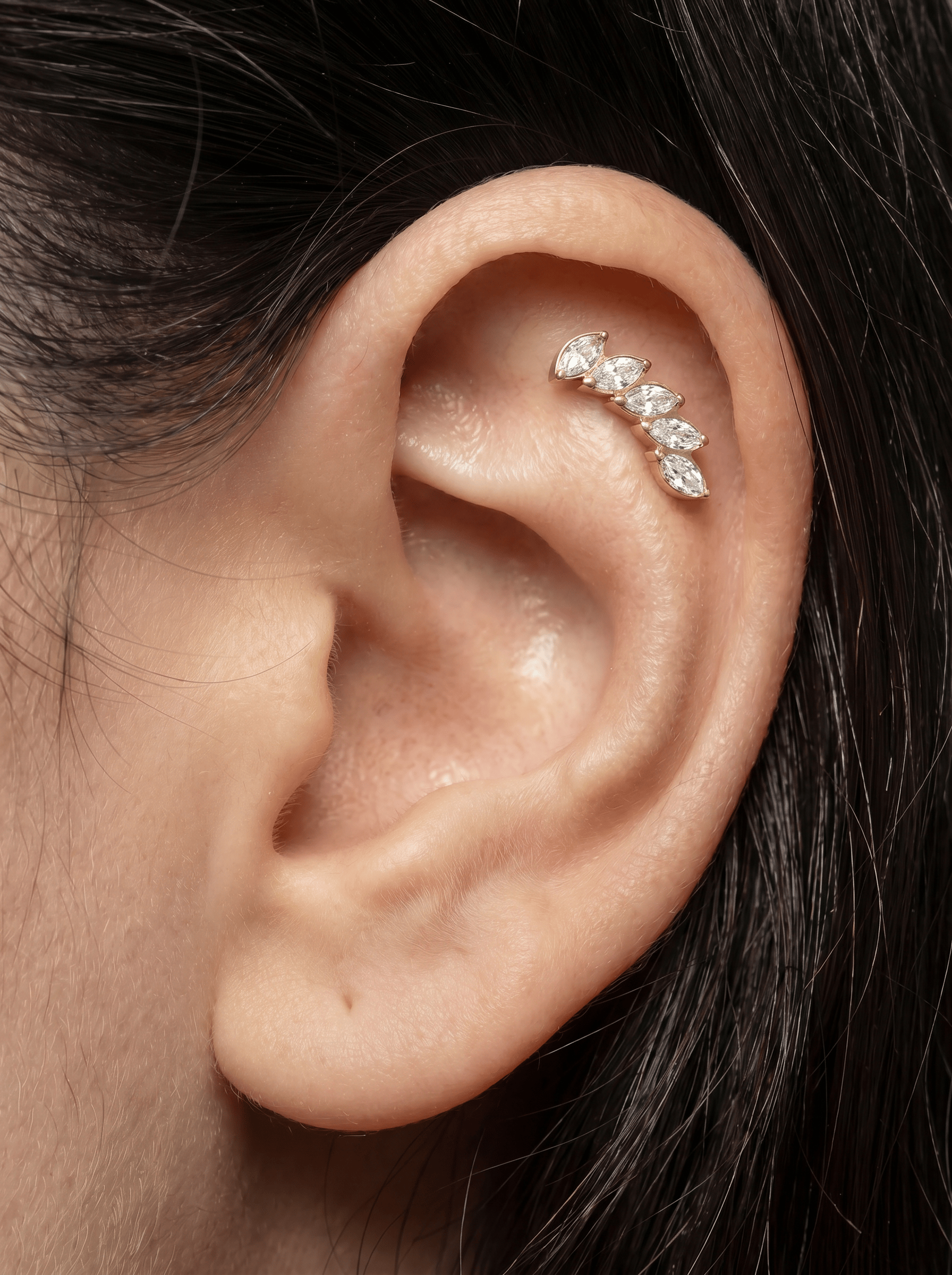 Un primer plano de la oreja de una persona con un piercing en el que se aprecia el pendiente de cartílago «Gia dorado» de Titanio-Noble Art, con cinco piedras de talla marquesa dispuestas en una línea curva a lo largo de la parte superior de la oreja.